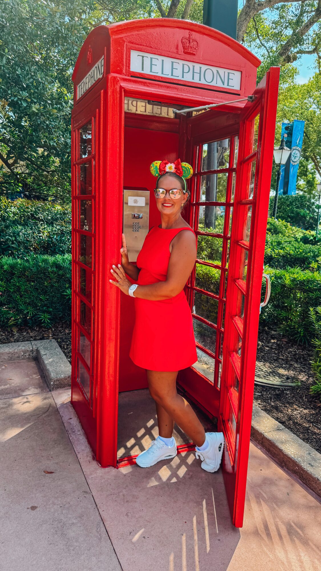 Person standing inside a red telephone booth in a leafy outdoor setting.