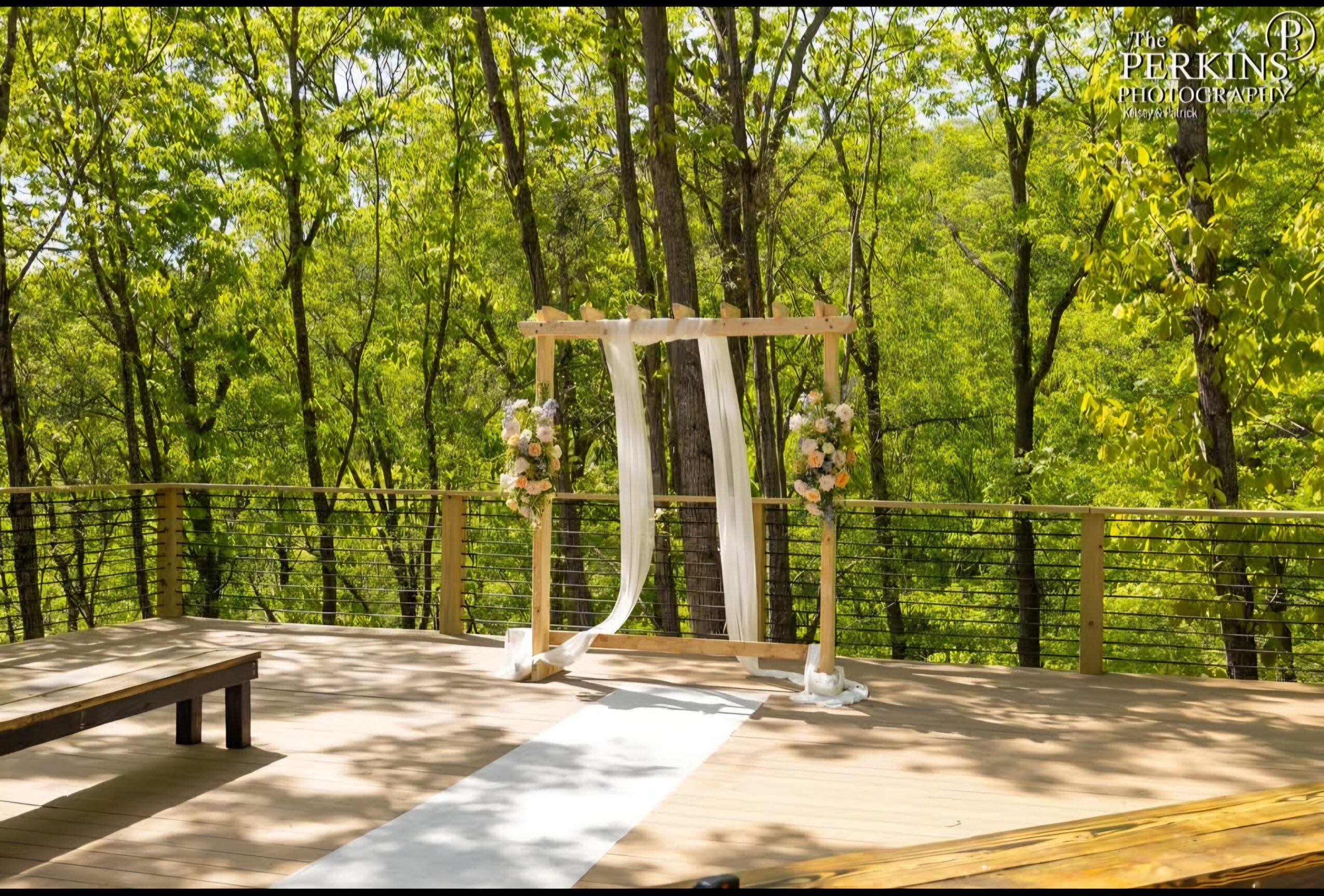 A wooden ceremony arch with white fabric stands on a sunlit deck surrounded by green trees and a black railing.