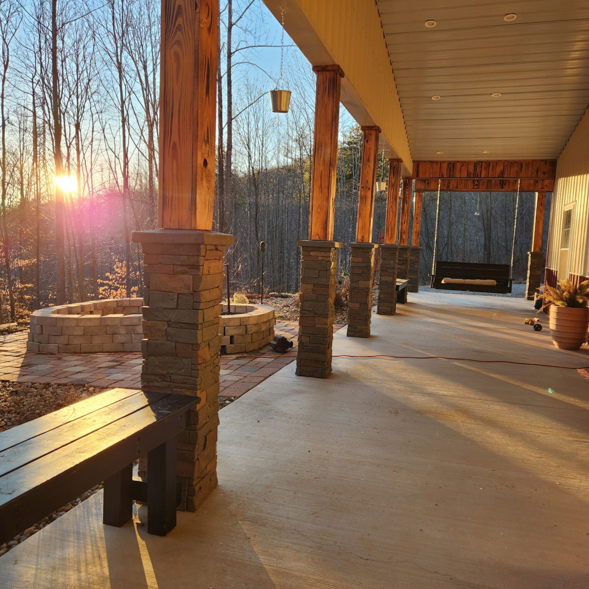 Covered wooden porch with stone-based columns, a bench, planters, and sunset light among bare trees.