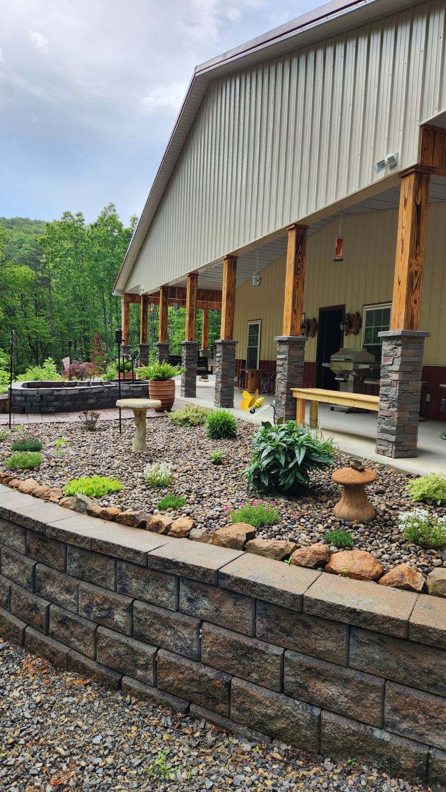 Covered outdoor patio beside a raised stone garden wall, with gravel, plants, seating, and trees.