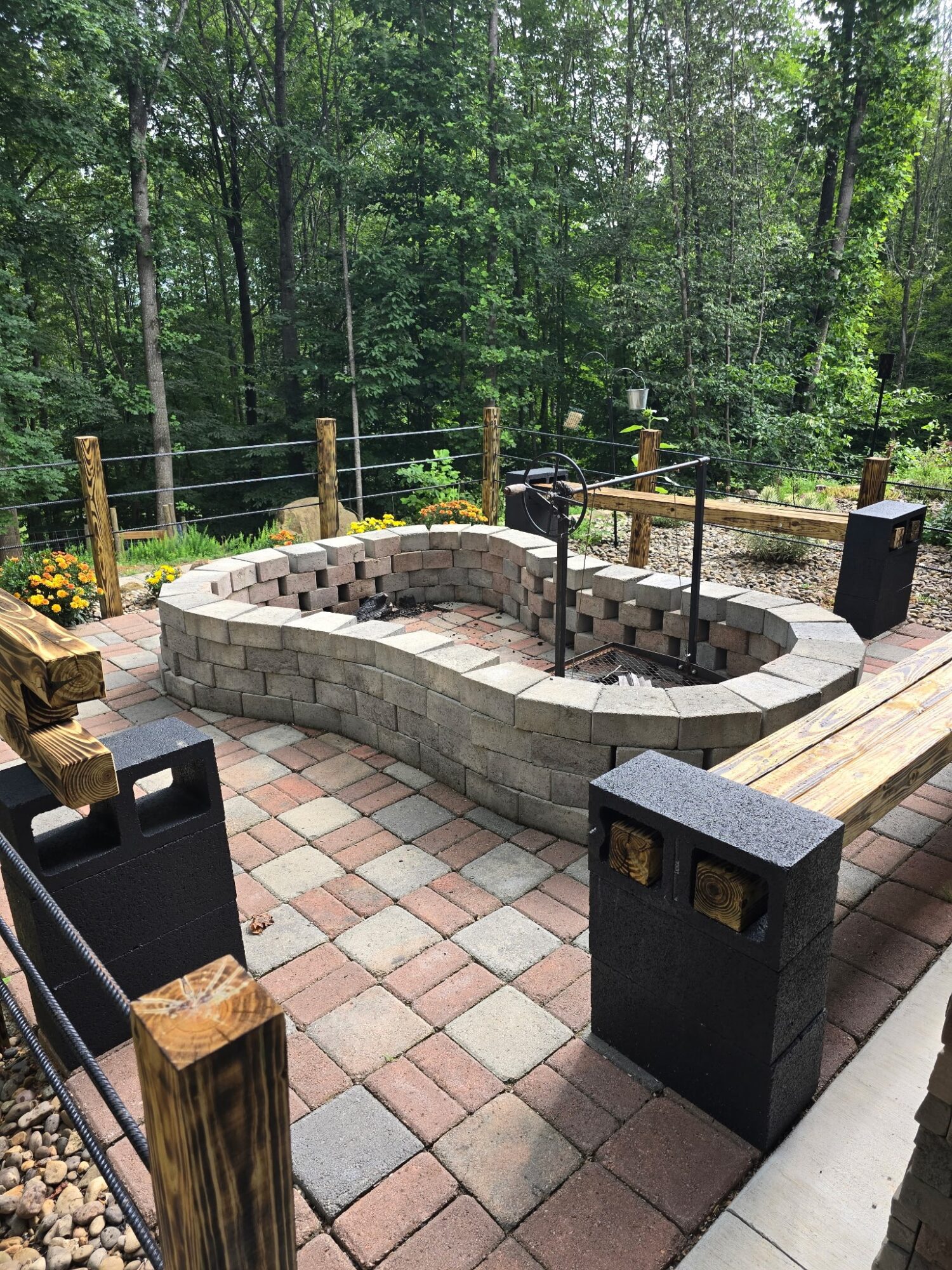 Stone fire pit surrounded by a paved patio, wooden benches, black posts, and trees.