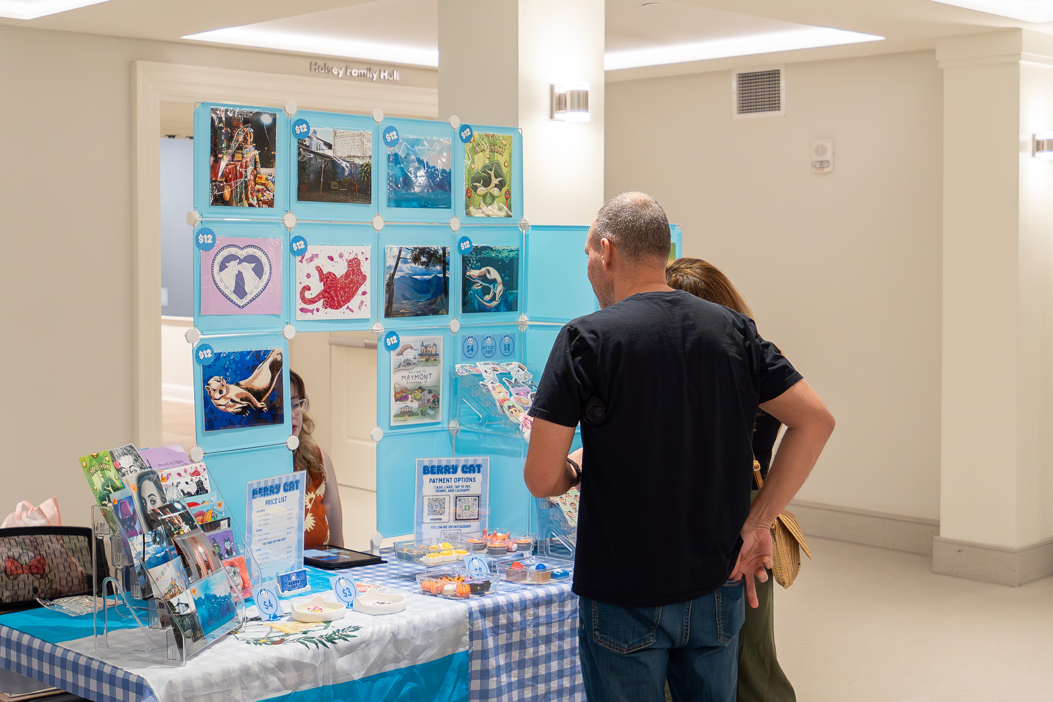Two people stand at a checkered table displaying books and colorful artwork in a bright room.