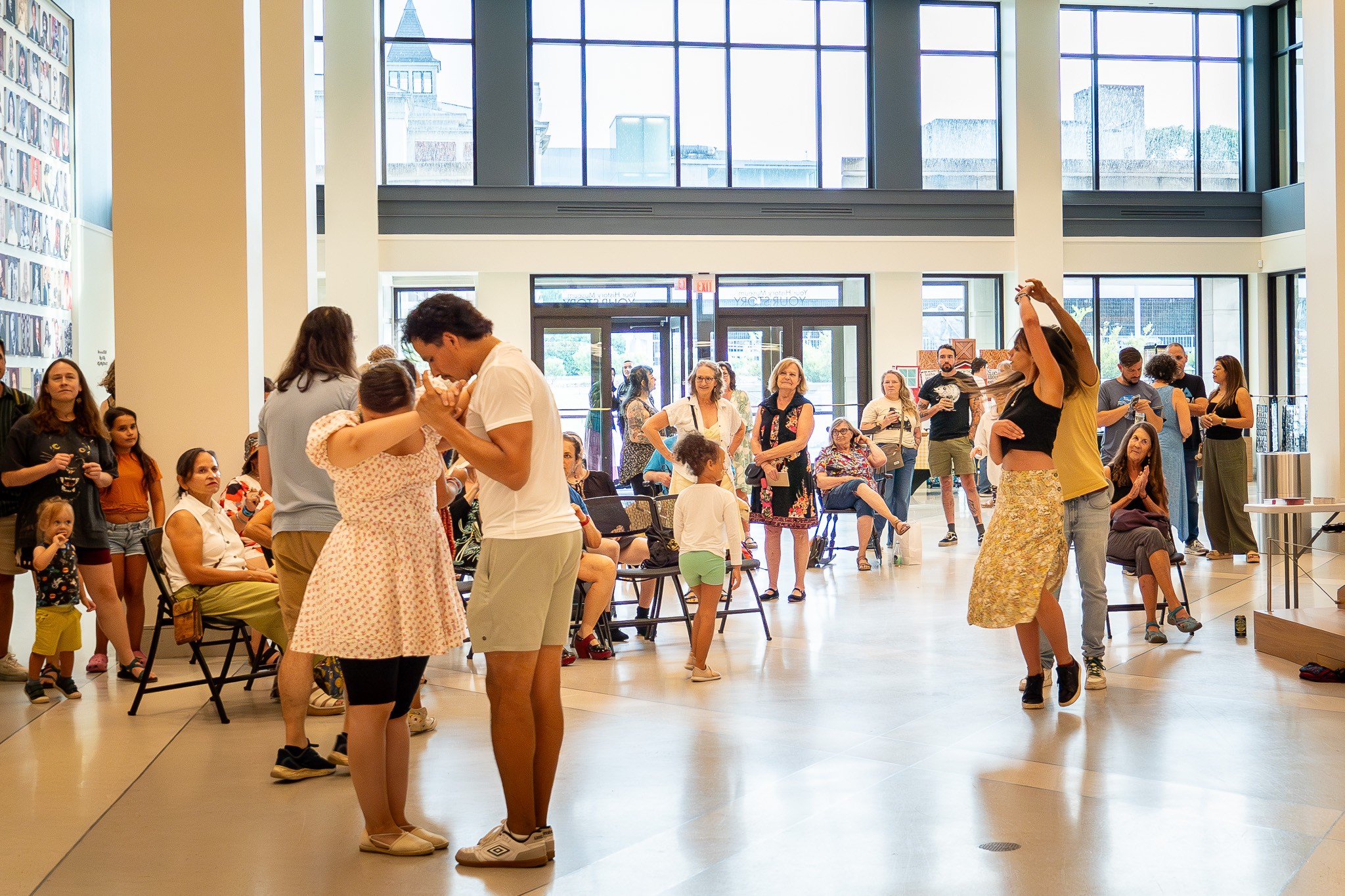 People gather and dance in a bright hall with large windows and chairs along the sides.