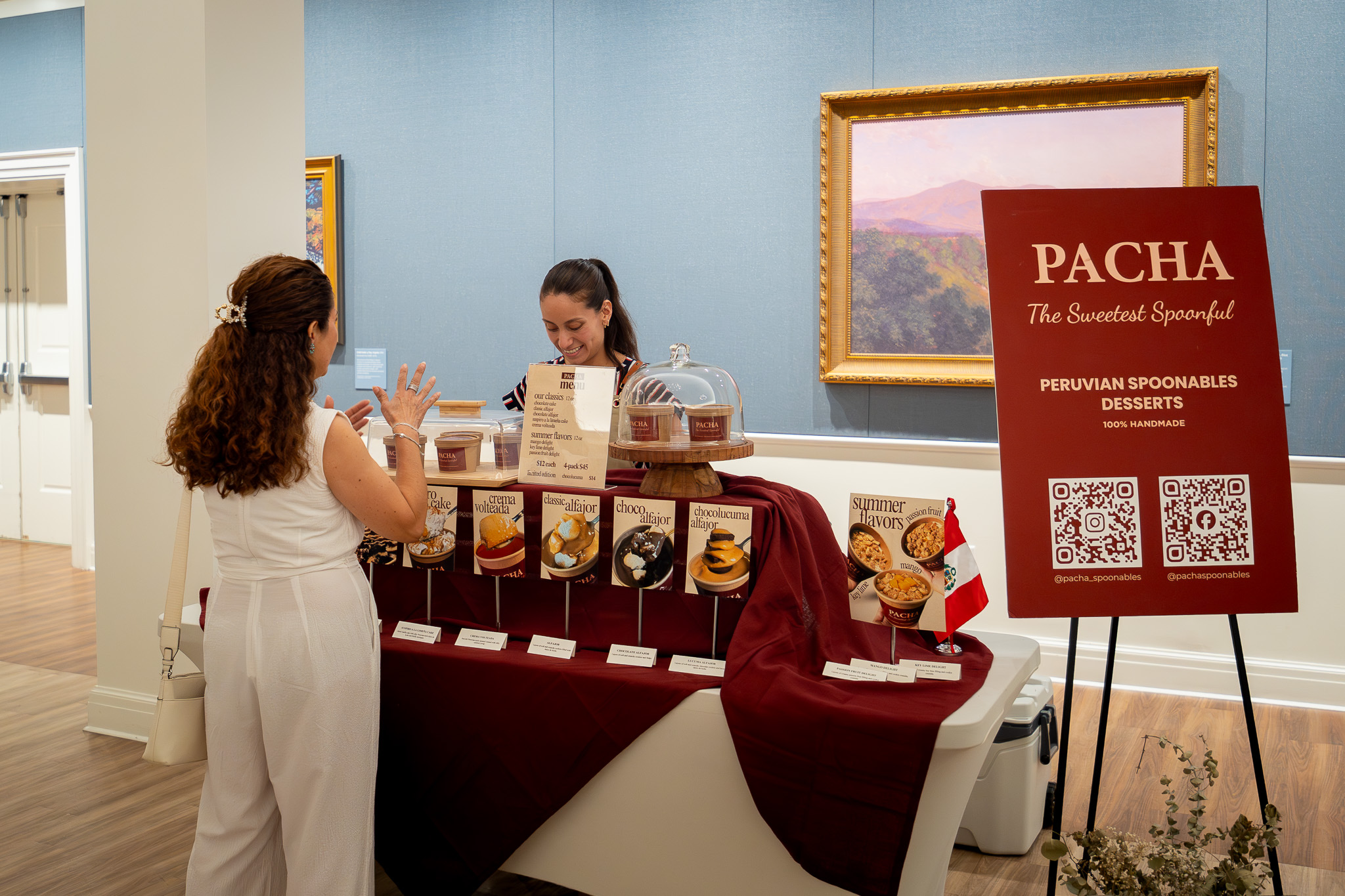 Two people stand at a dessert display beside a red Pacha sign with QR codes.