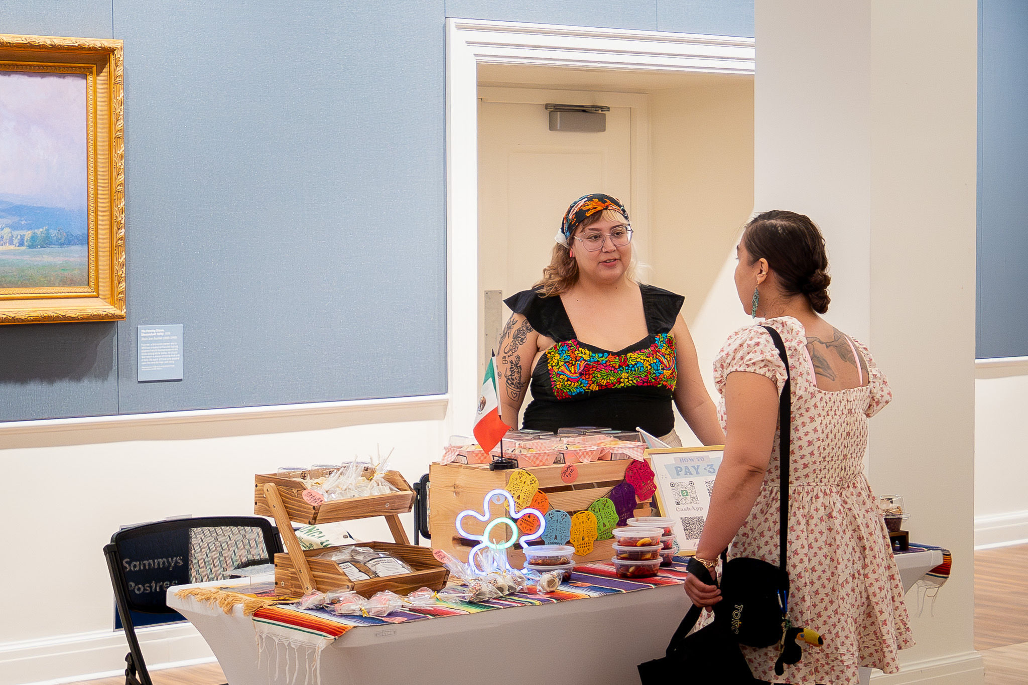 Two people stand beside a table displaying colorful handmade crafts in a gallery room.