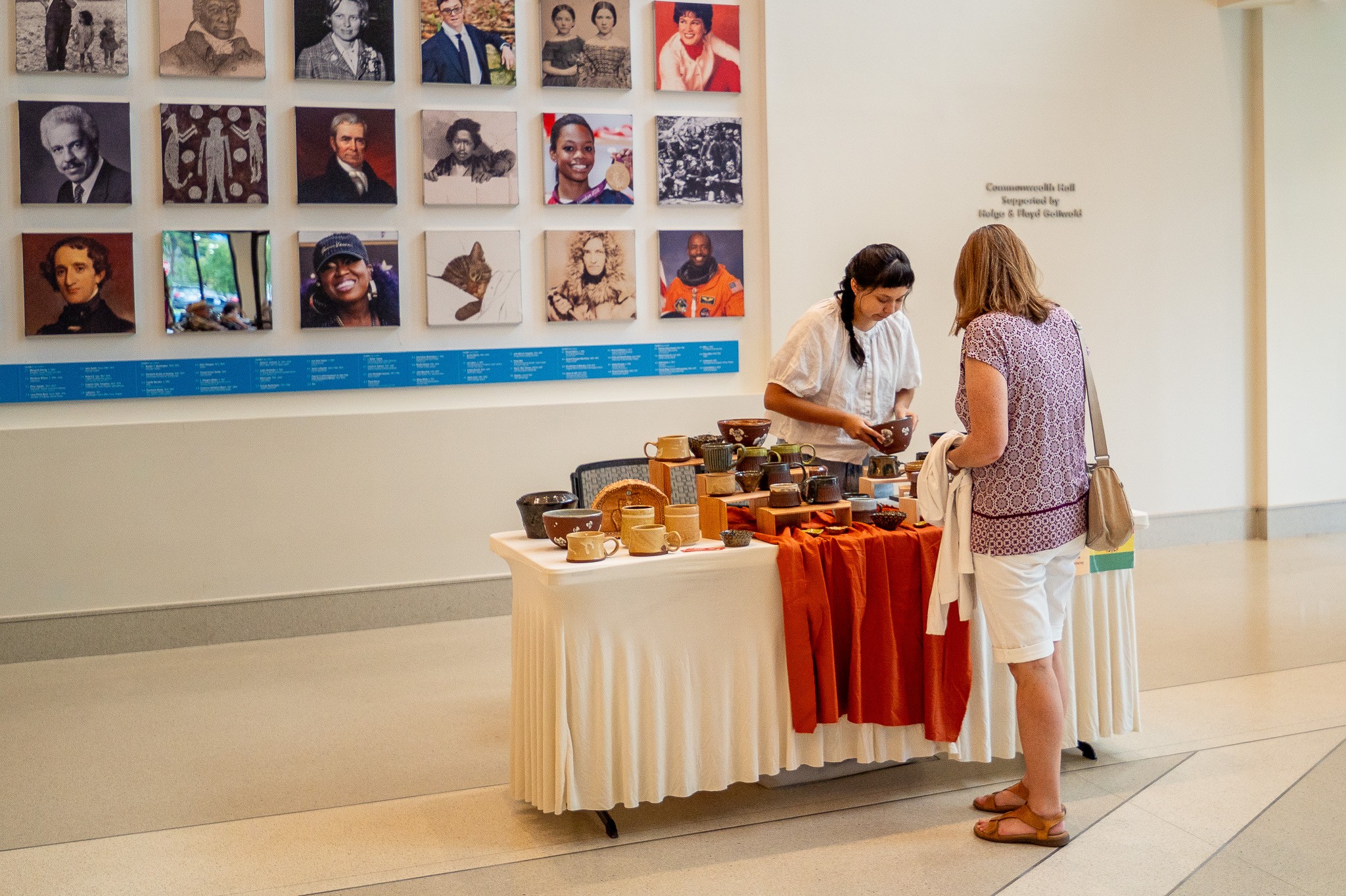 Two people stand beside a table displaying pottery in a gallery with a wall of portrait photographs.