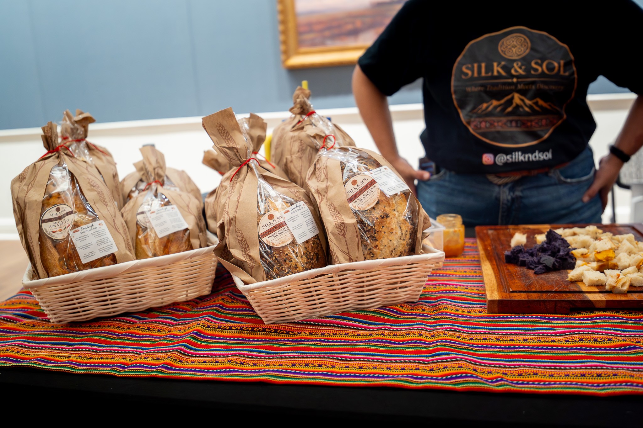 Wrapped baked goods in baskets on a colorful tablecloth beside a person and serving plates.