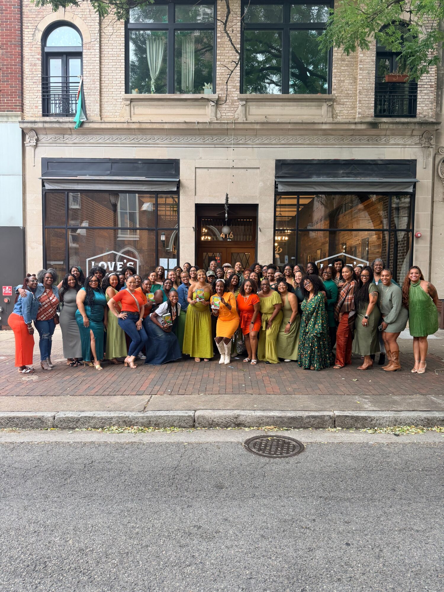 A group of people stands along a sidewalk in front of a brick building.