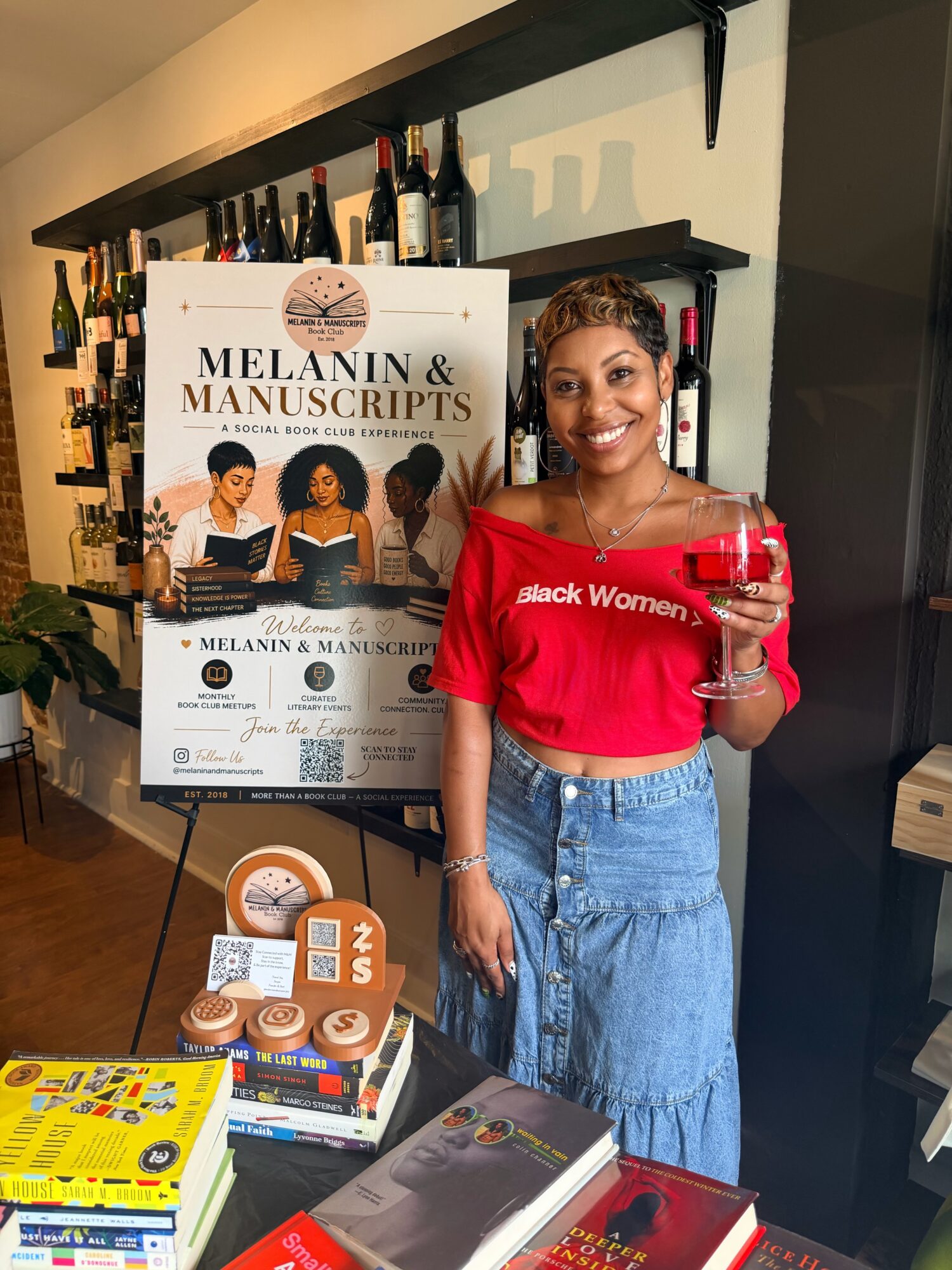 Person in a red shirt holds a glass beside a table of books and a Melanin & Manuscripts display.
