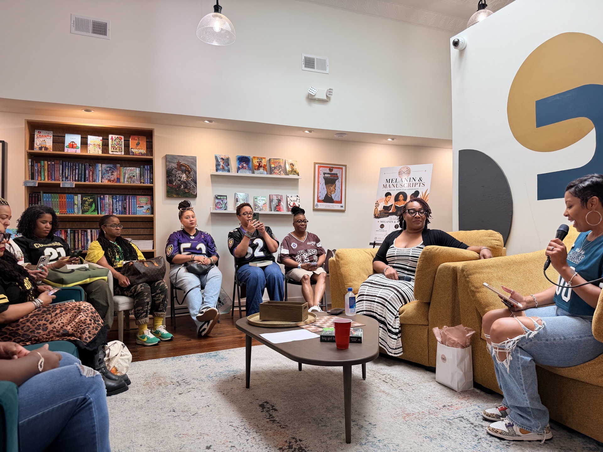 People sit in a living room around a coffee table, with bookshelves and a large abstract wall decoration behind them.