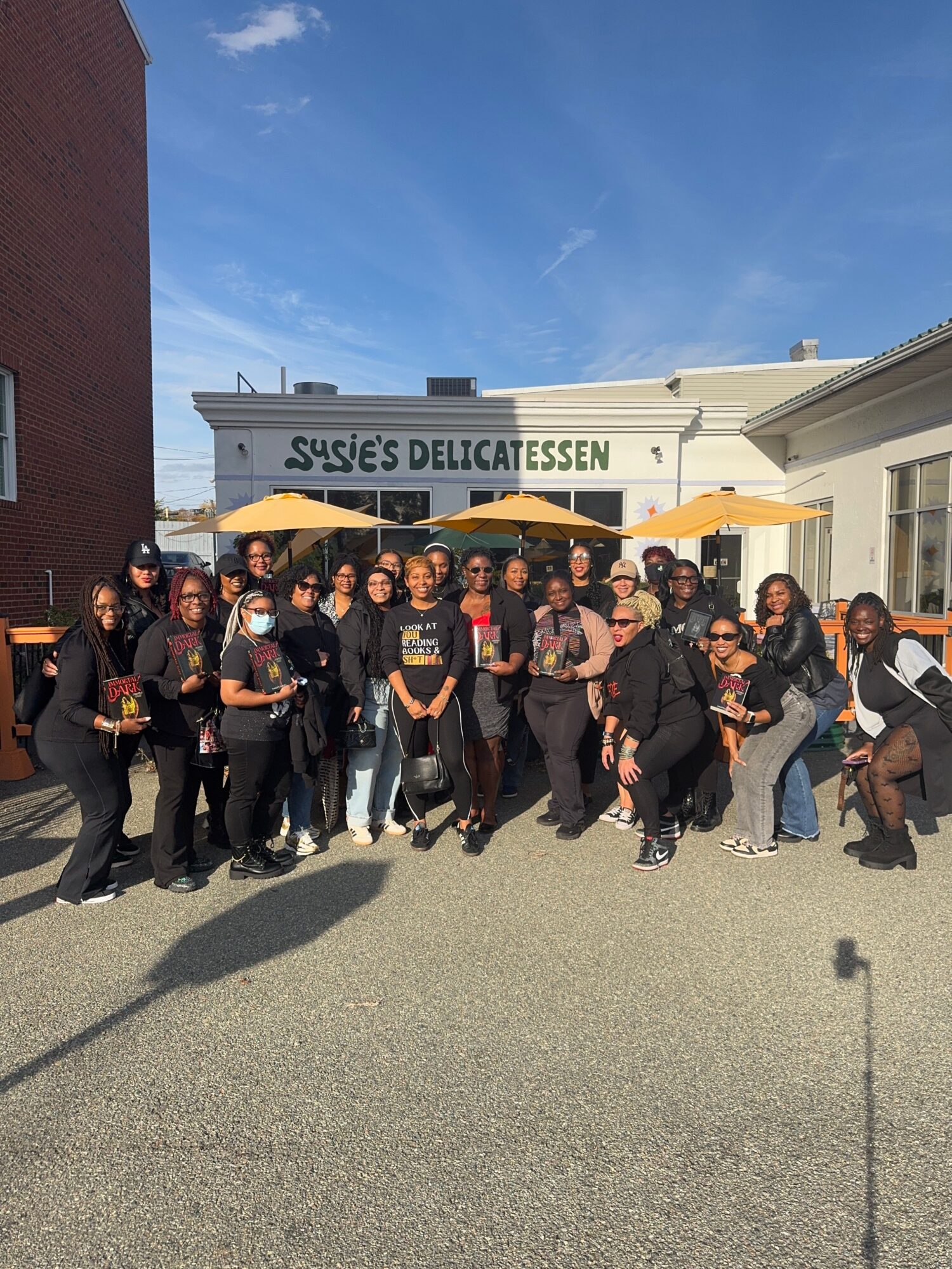 A group of people pose outside a deli beneath an awning, with a brick wall and blue sky nearby.