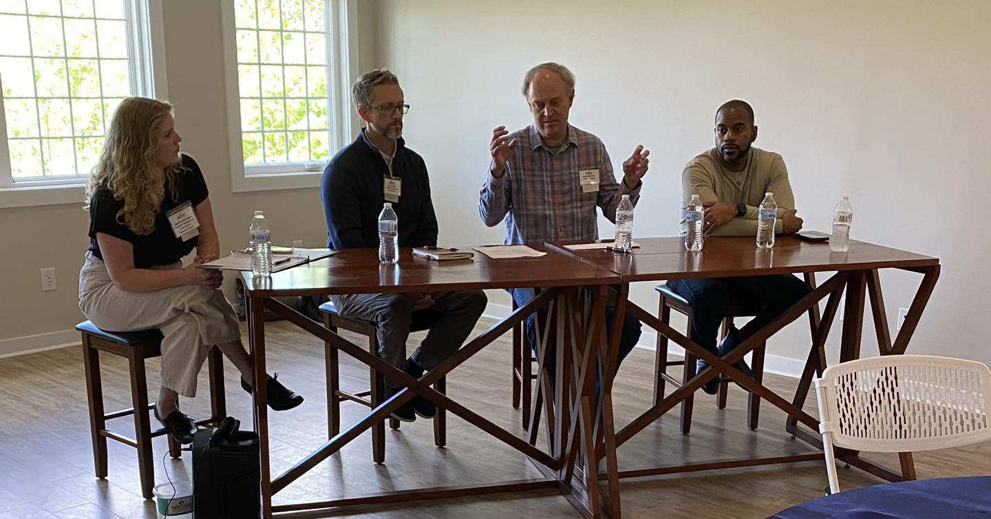 Four people sit at a long table with water bottles and papers in a bright room.