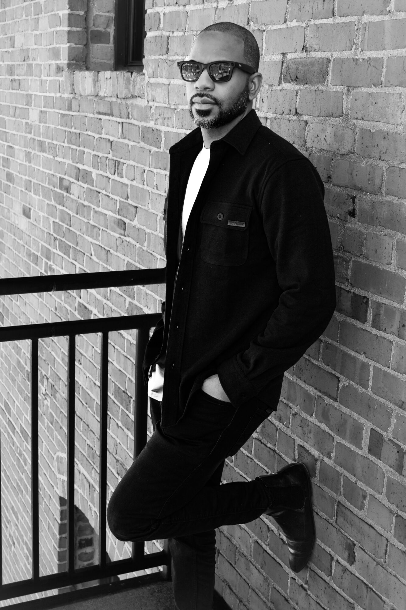 Person wearing sunglasses and a dark coat leans against a brick wall beside a metal railing.