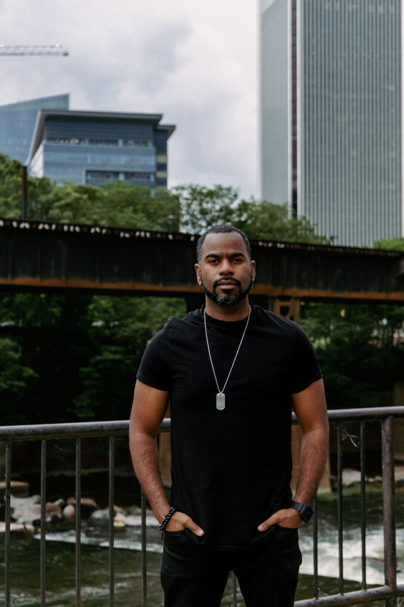 A person in a black shirt stands by a railing, with trees and tall buildings behind him.
