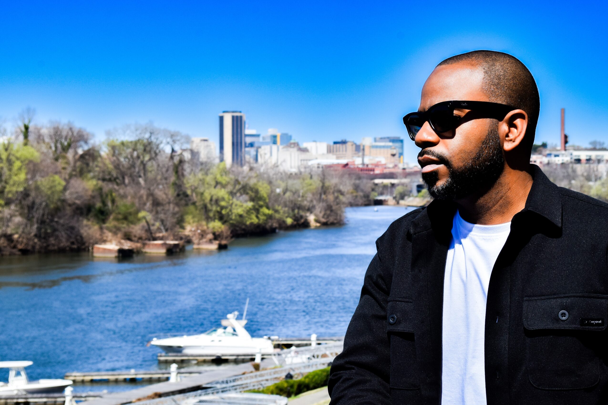 A person wearing sunglasses and a black jacket stands beside a river, with boats and a city skyline behind him.