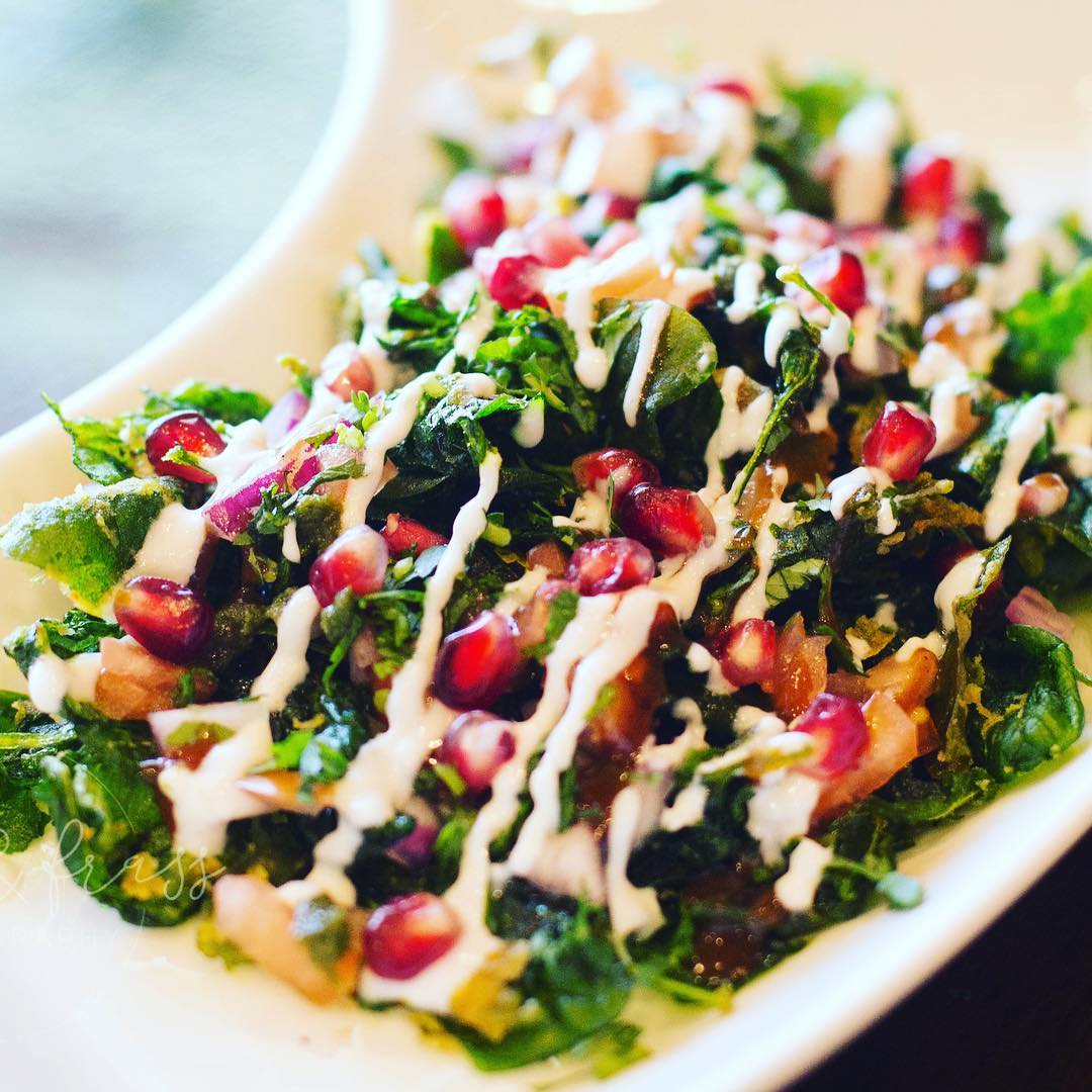 A leafy salad topped with pomegranate seeds, chopped vegetables, and creamy dressing on a white plate.