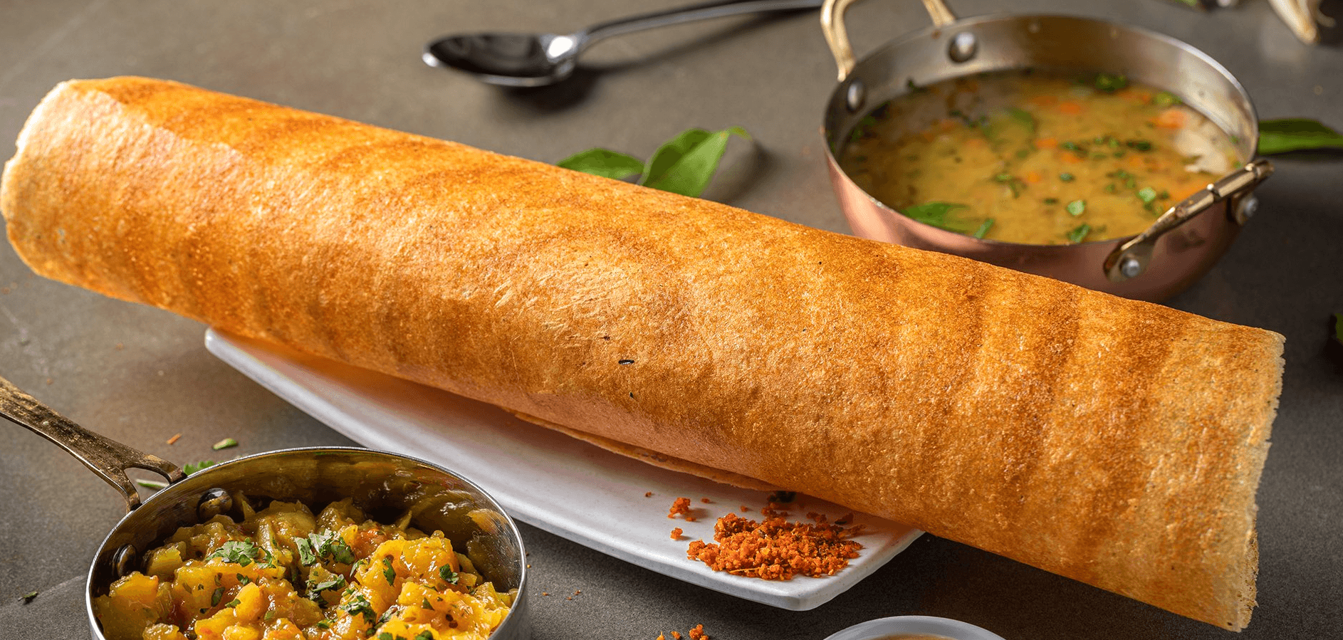 Long golden-brown crepe on a platter beside bowls of curry and chutney.