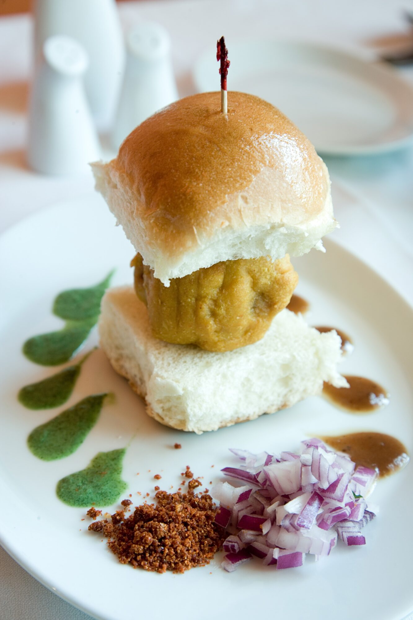 A plated appetizer with a bun, layered filling, chopped purple onion, brown seasoning, and green sauce drops.