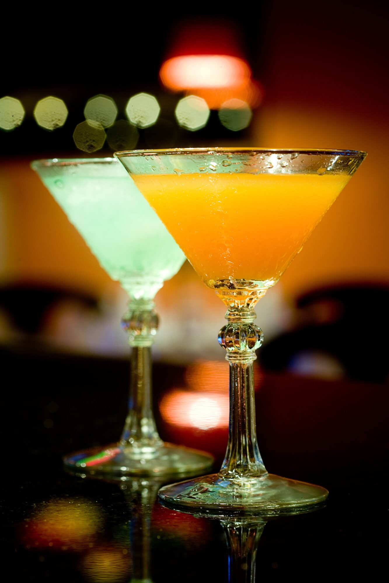 Two martini glasses filled with colorful drinks sit on a reflective bar beneath blurred lights.
