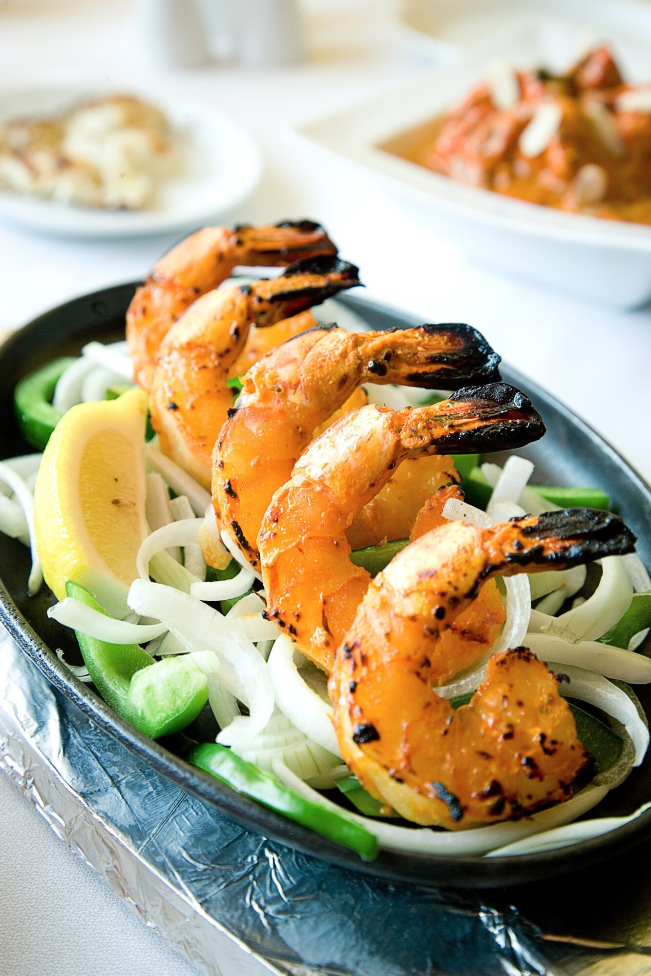 Grilled shrimp arranged over sliced vegetables and greens on an oval platter, with plates of food in the background.