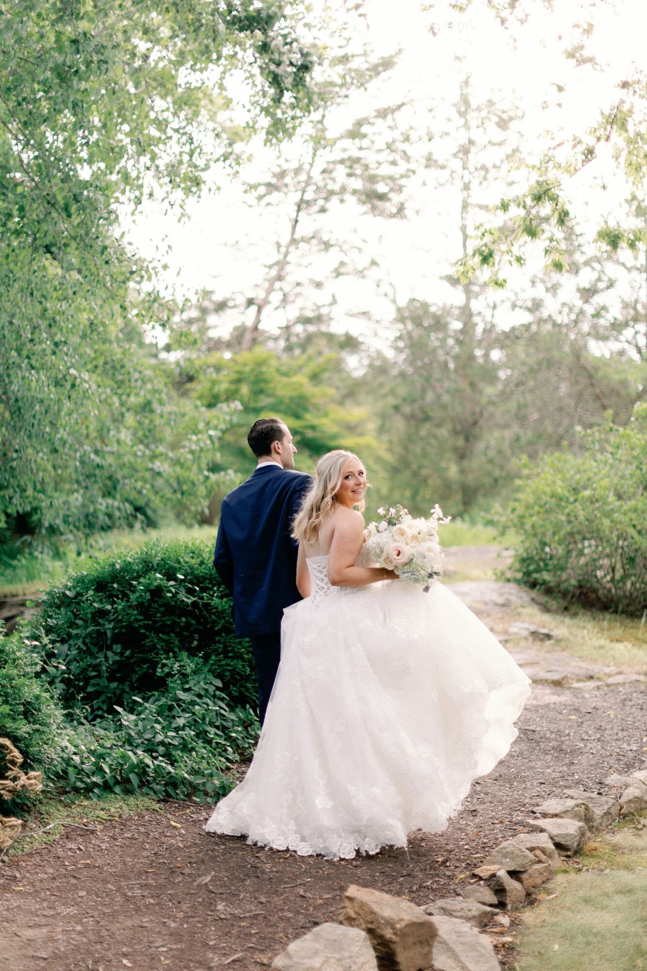 A couple stands on a woodland path, with one person wearing a flowing white gown and holding a bouquet.