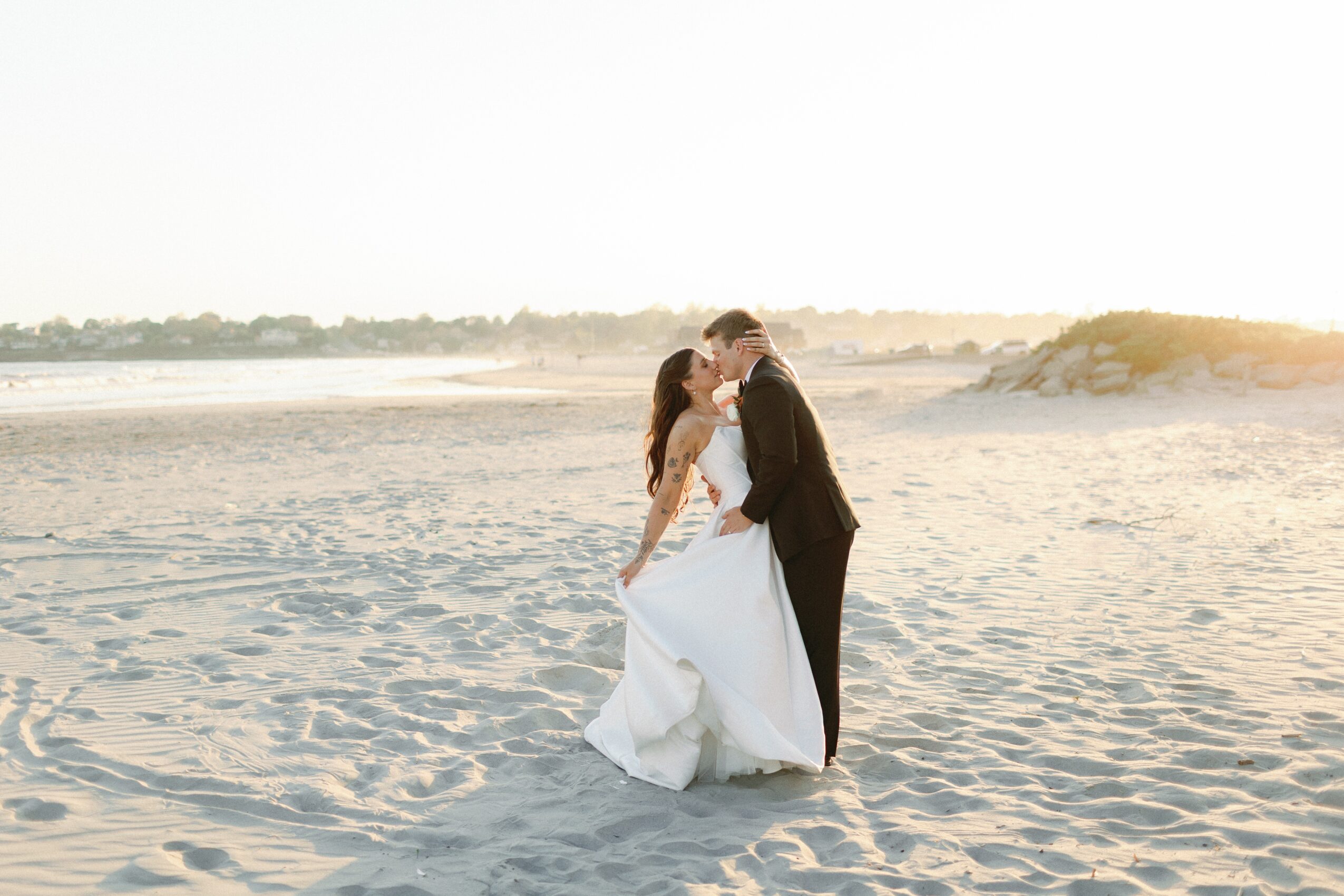 A couple embraces on a sandy beach beside the water, with dunes in the background.