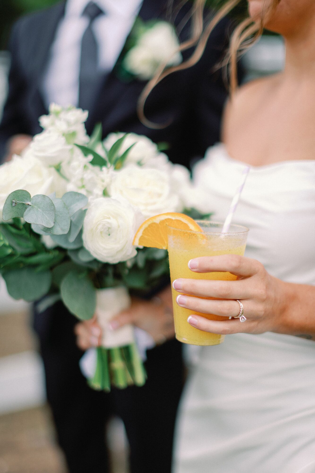 A person in a white dress holds a yellow drink beside a bouquet, with another person in a dark suit behind them.