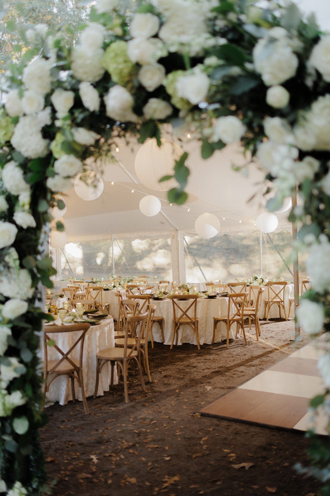 Floral arch framing a decorated event tent with long tables, chairs, and a dance floor.