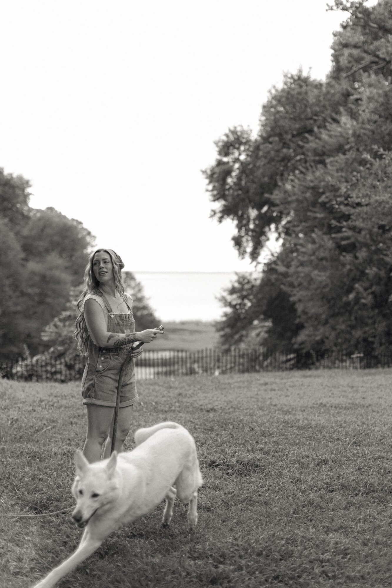 A person stands on a lawn beside a large white dog, with trees and a body of water behind them.