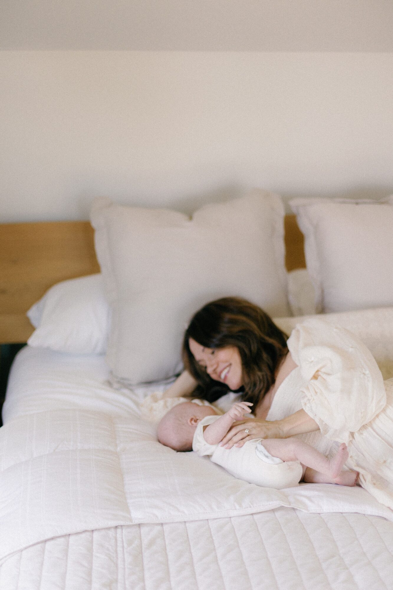 A person sitting on a bed cradles a baby beside white pillows and bedding.