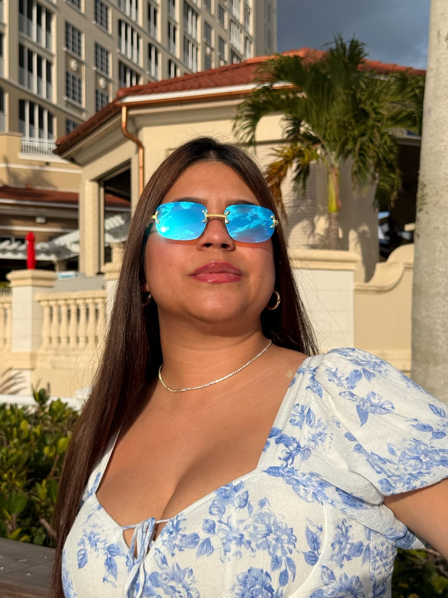 Person wearing reflective blue sunglasses and a floral dress outdoors beside palm trees and buildings.
