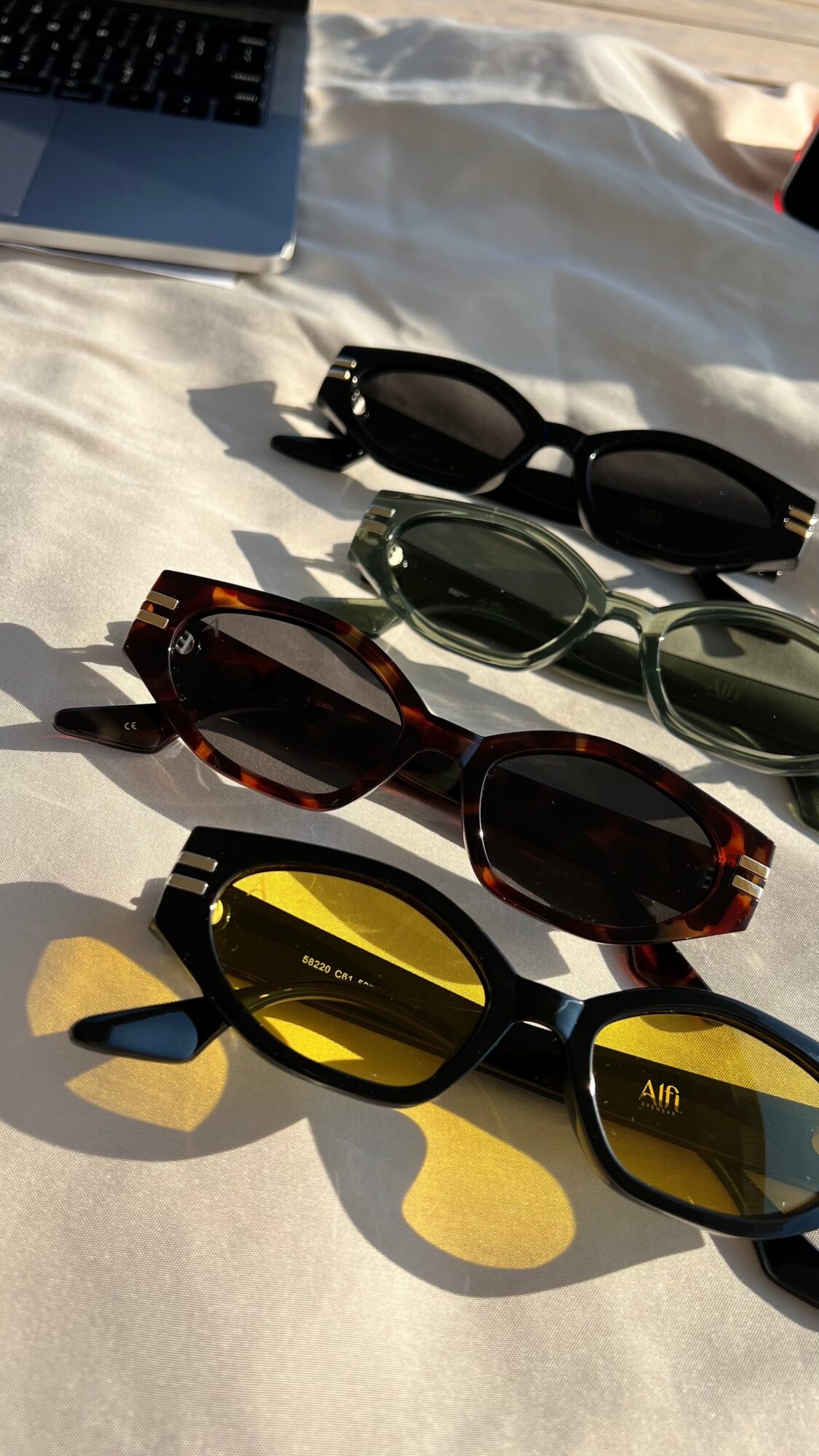 Four pairs of sunglasses lie on a light fabric surface beside part of a laptop.