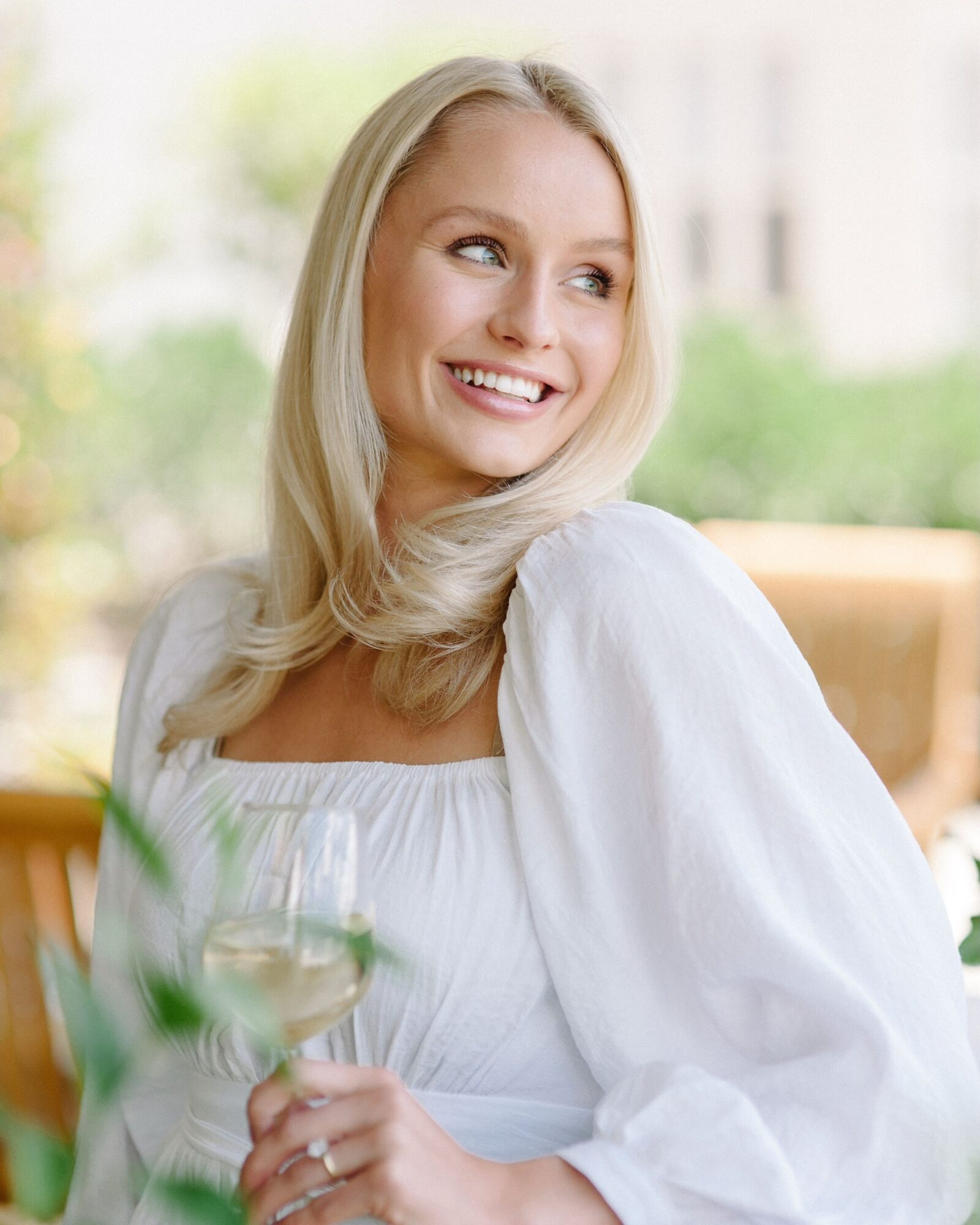Person with long blonde hair wearing a white dress, holding a glass near a table.