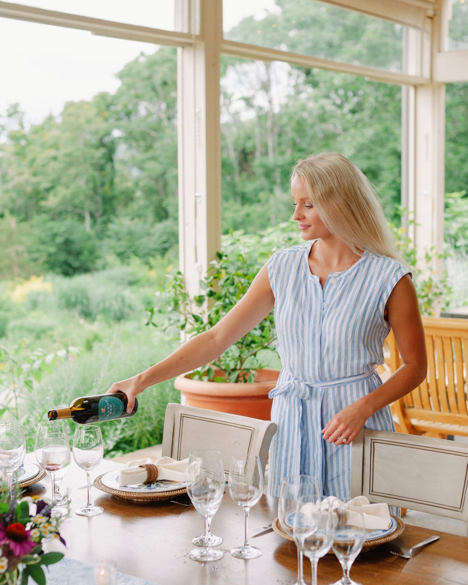 A woman reaches toward a bottle beside a dining table set with plates and glasses, near a window overlooking greenery.