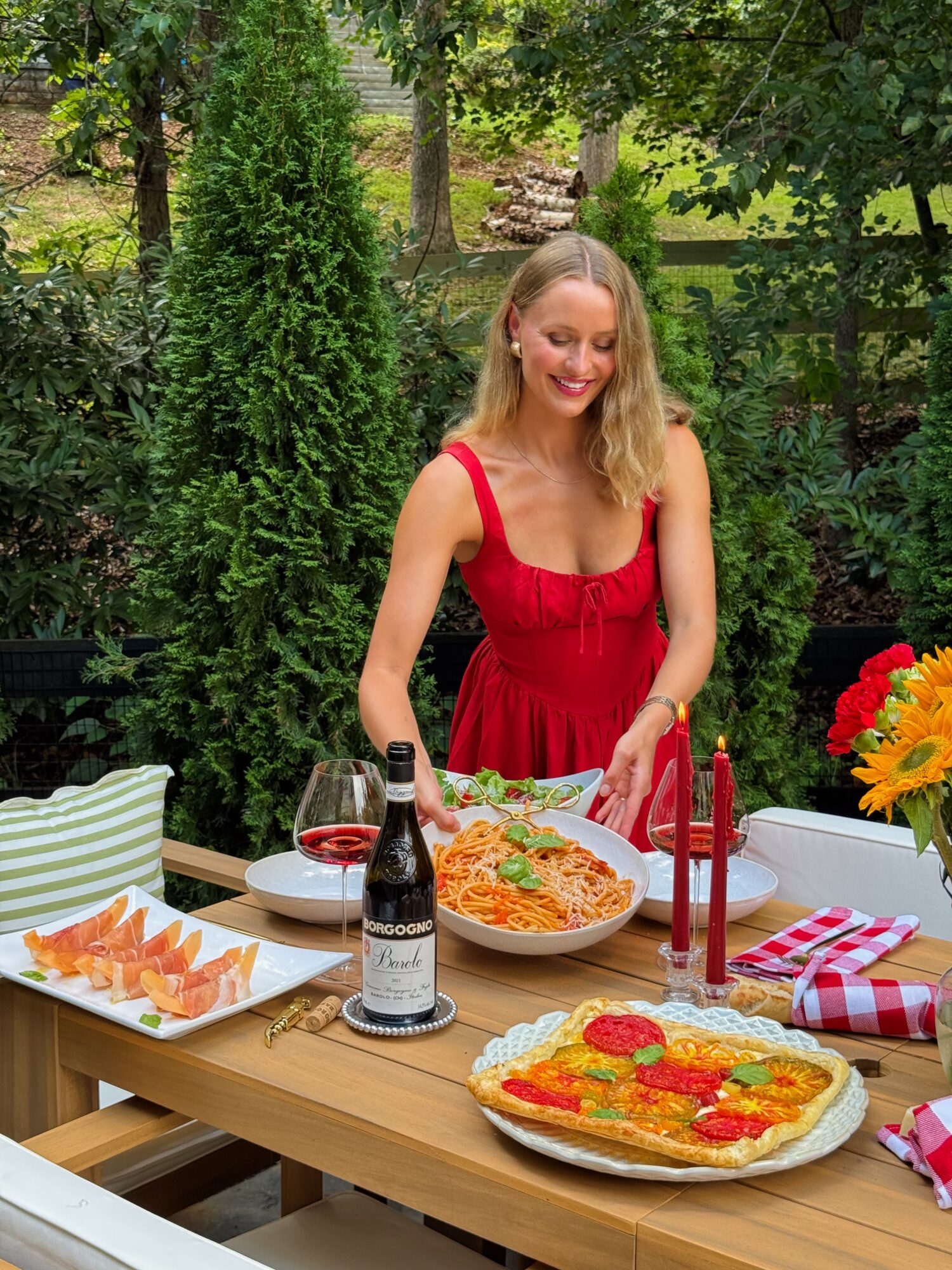 A woman in a red dress serves pasta at an outdoor table set with food and flowers.