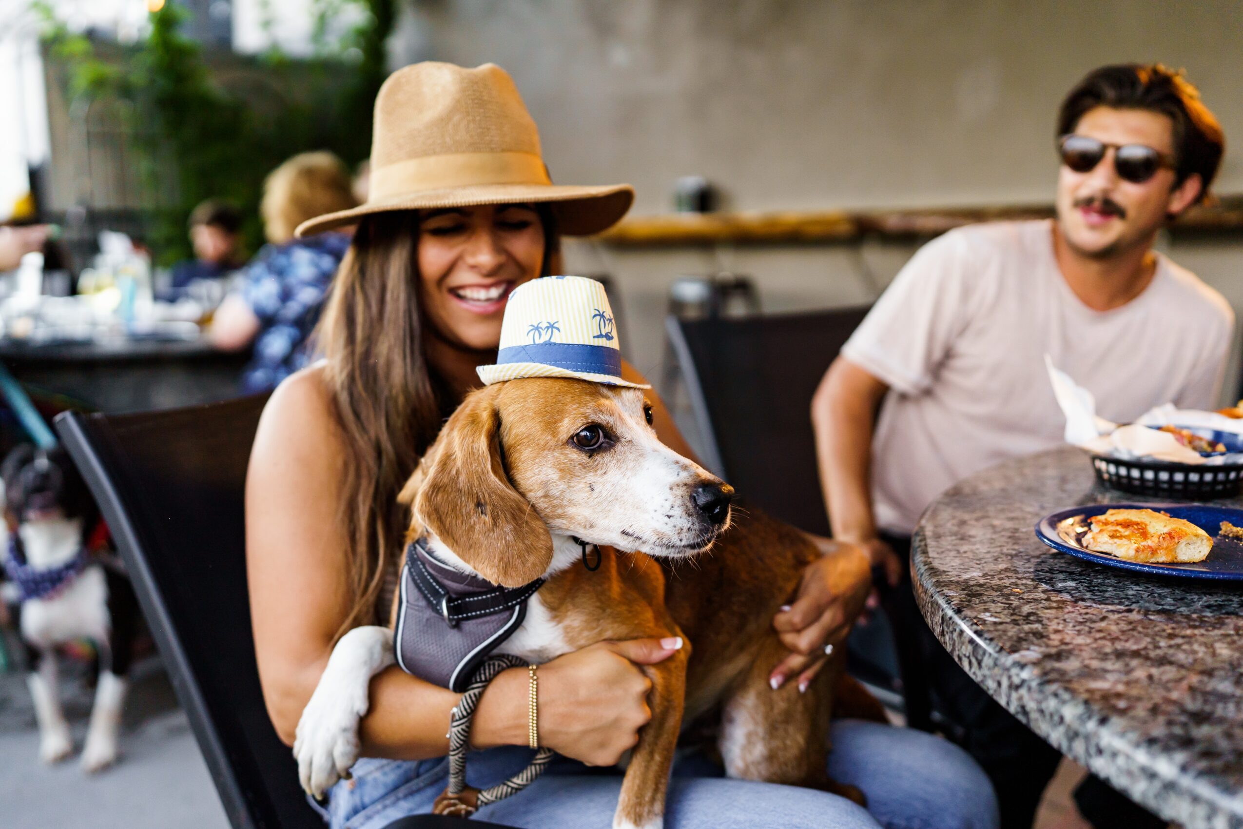 A woman in a straw hat holds a dog wearing a blue-and-white hat at an outdoor café, with a man seated nearby.