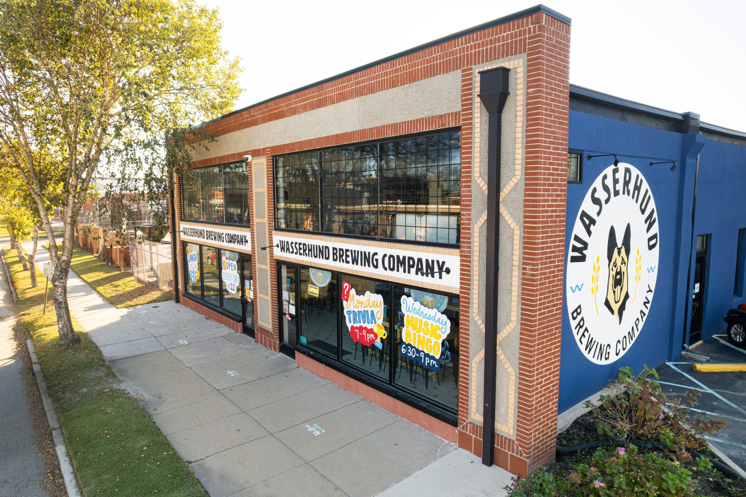 Brick-and-blue brewery building with large windows, signage, and a sidewalk alongside it.