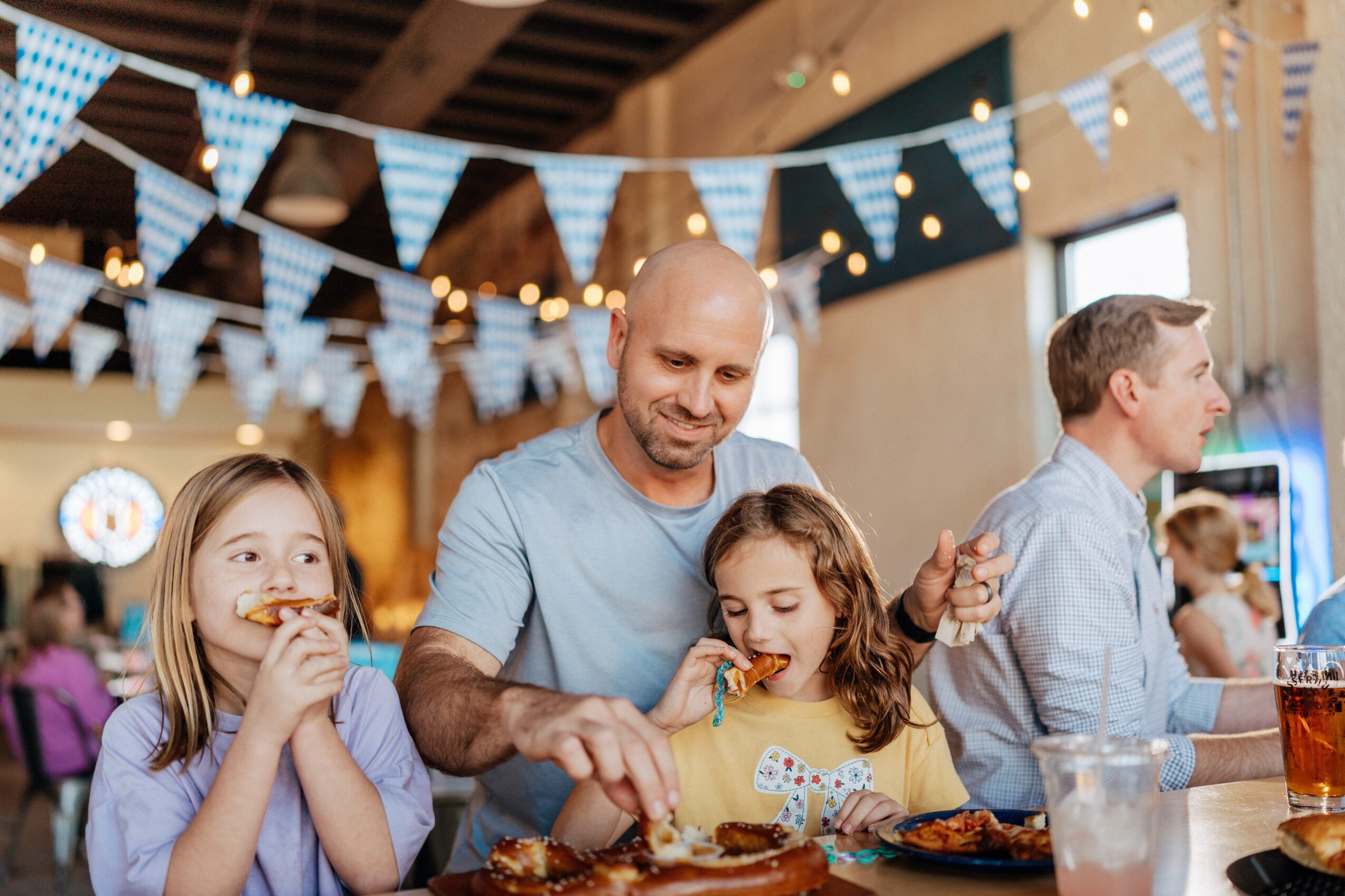 A man and two children stand at a buffet table beneath bunting and string lights, with other people nearby.