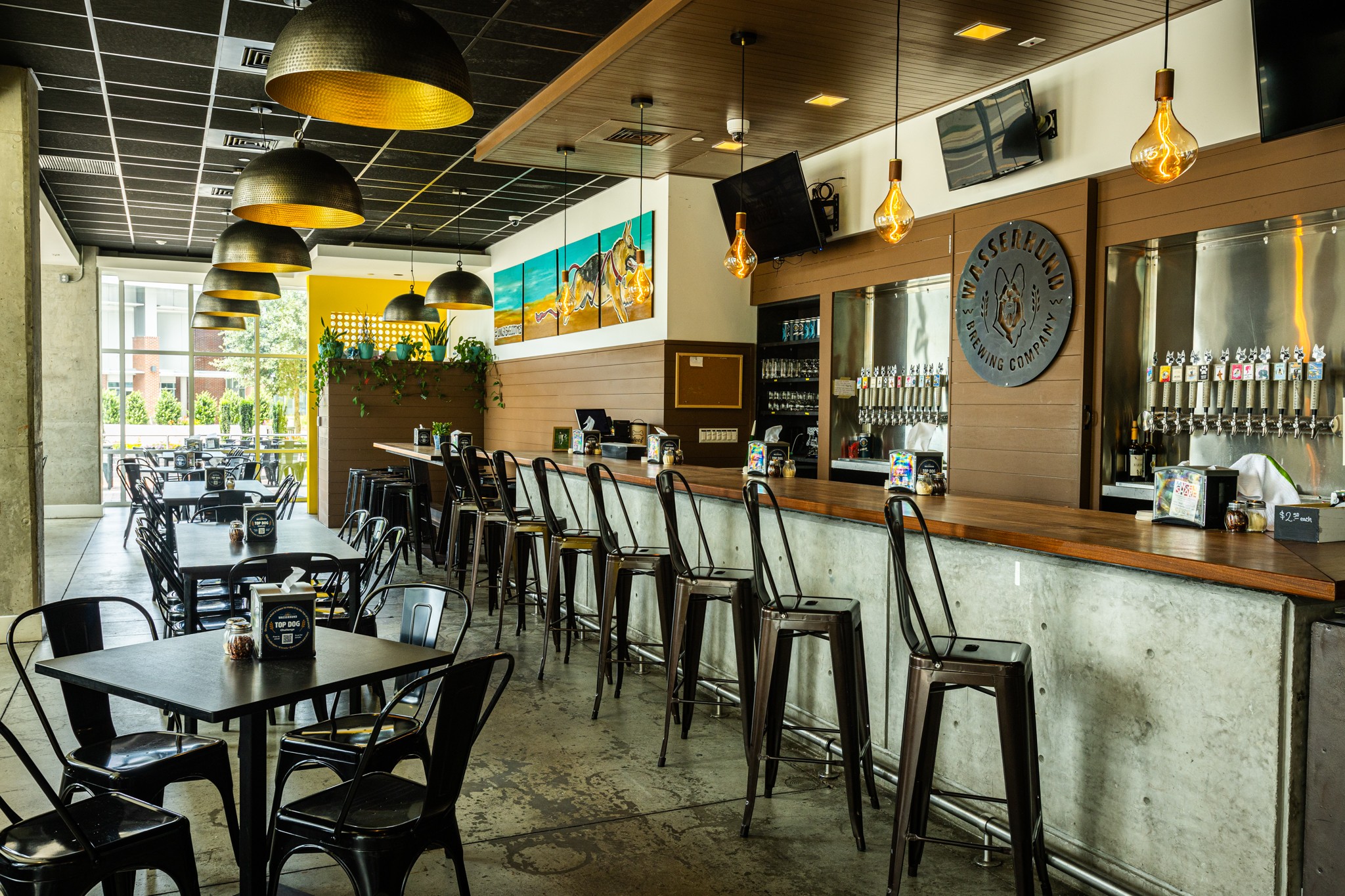 Interior of a restaurant bar with a counter, stools, tables, pendant lights, and large windows.