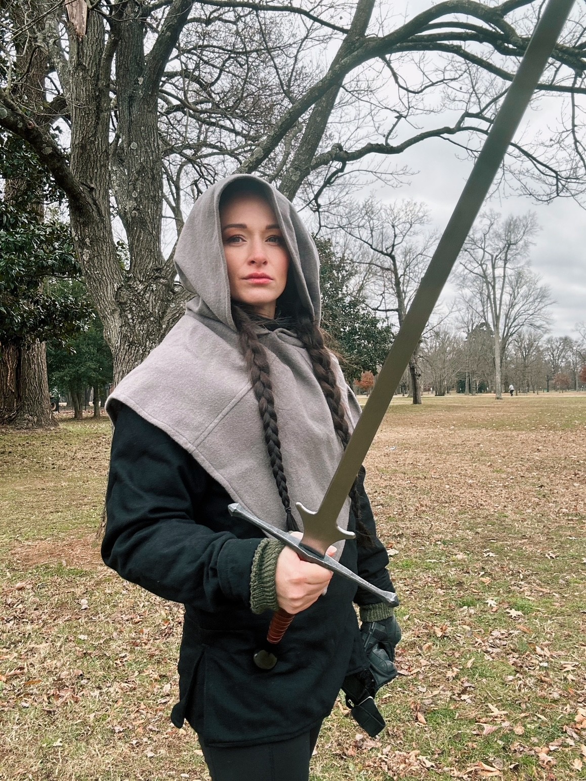 Person wearing a gray hooded cloak holds a sword in a leaf-covered park beneath bare trees.