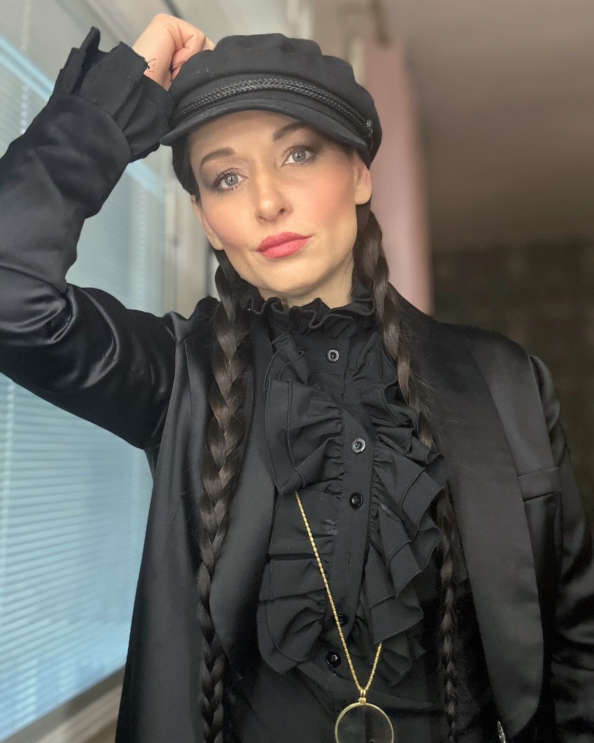 Person wearing a black cap and outfit, with two braids, raising a hand to the cap beside a window.