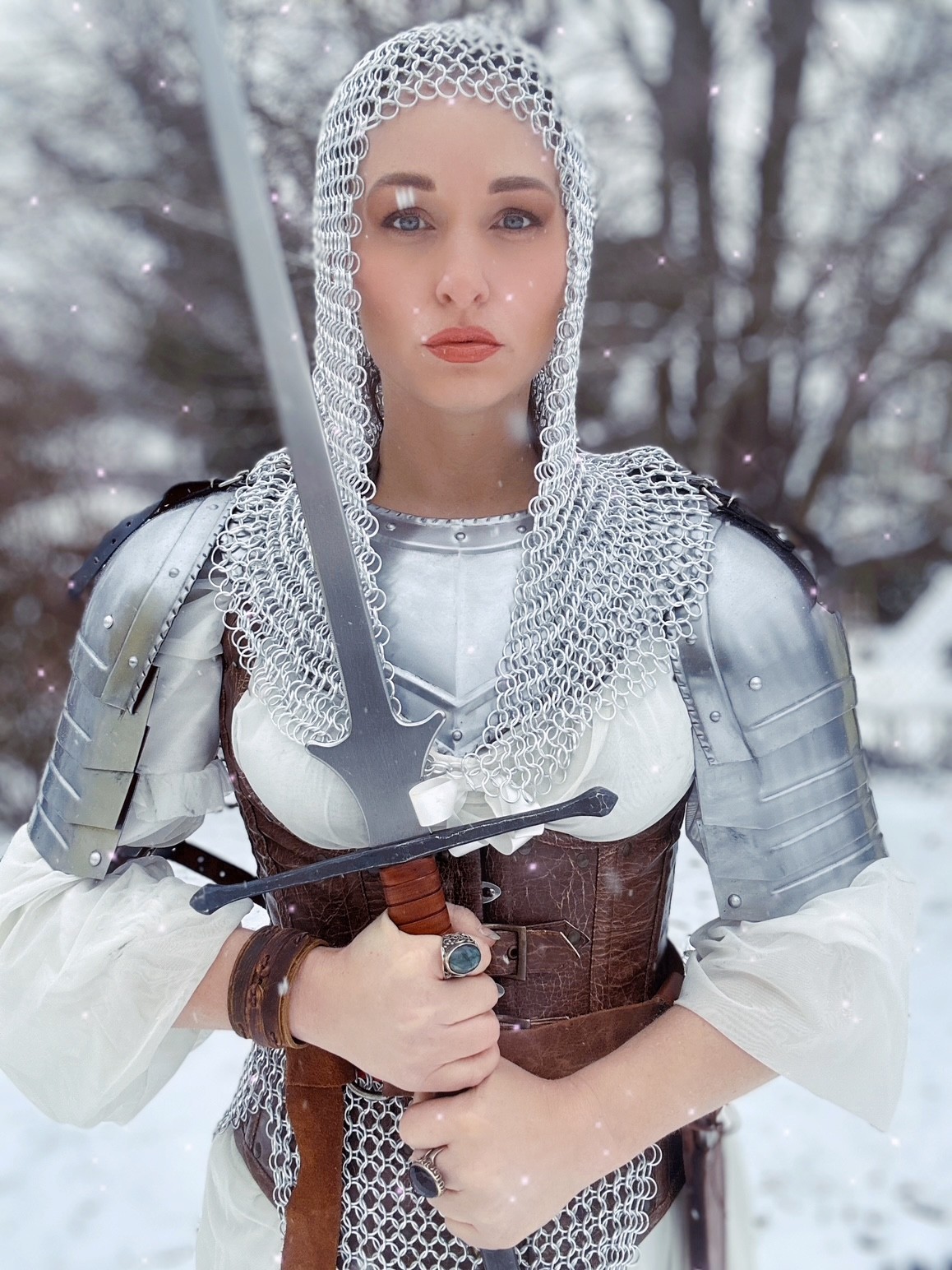 Person wearing chainmail and plate armor stands in a snowy forest, holding a sword.