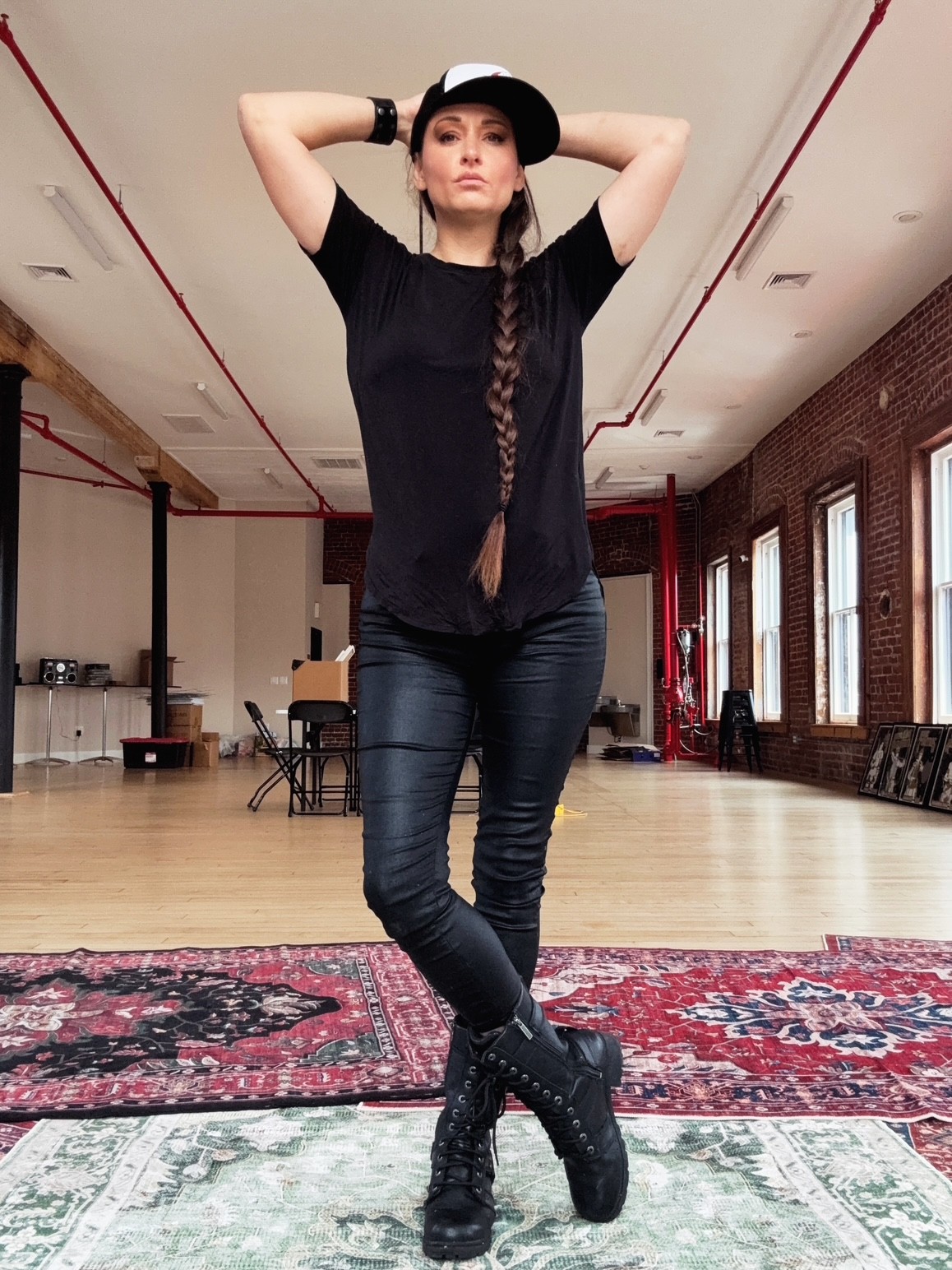 Person in a black hat stands with crossed legs on patterned rugs in a long indoor hall.