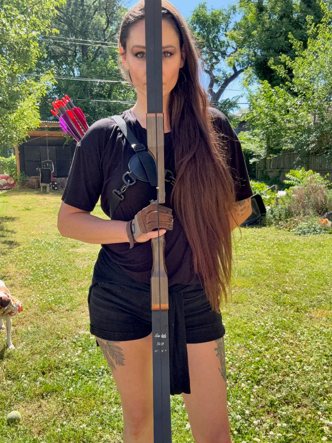 A person holding a bow stands on grass outdoors, with a quiver of arrows and trees nearby.