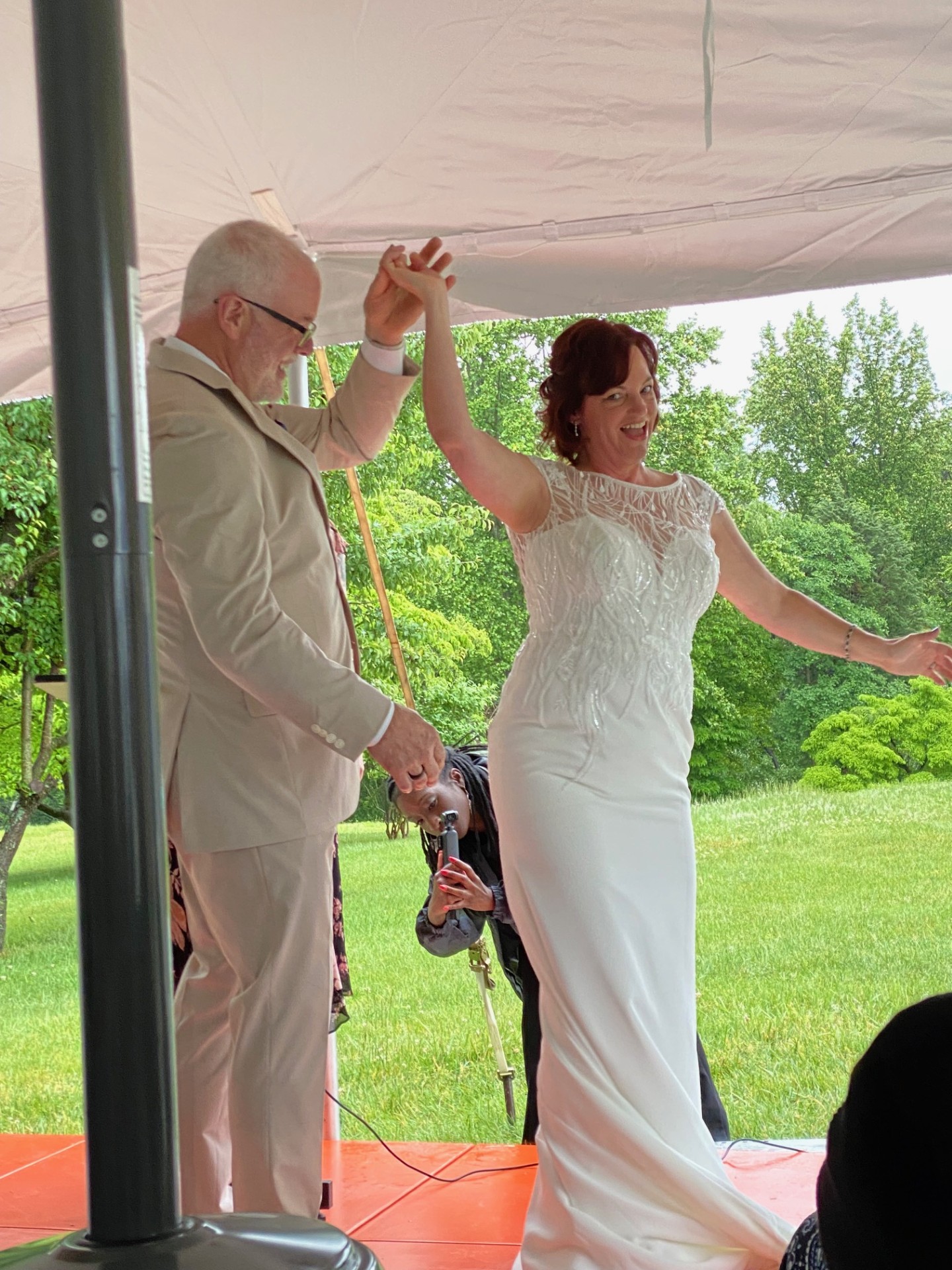 A person in a white dress dances with an older person beneath an outdoor canopy, with guests and greenery nearby.