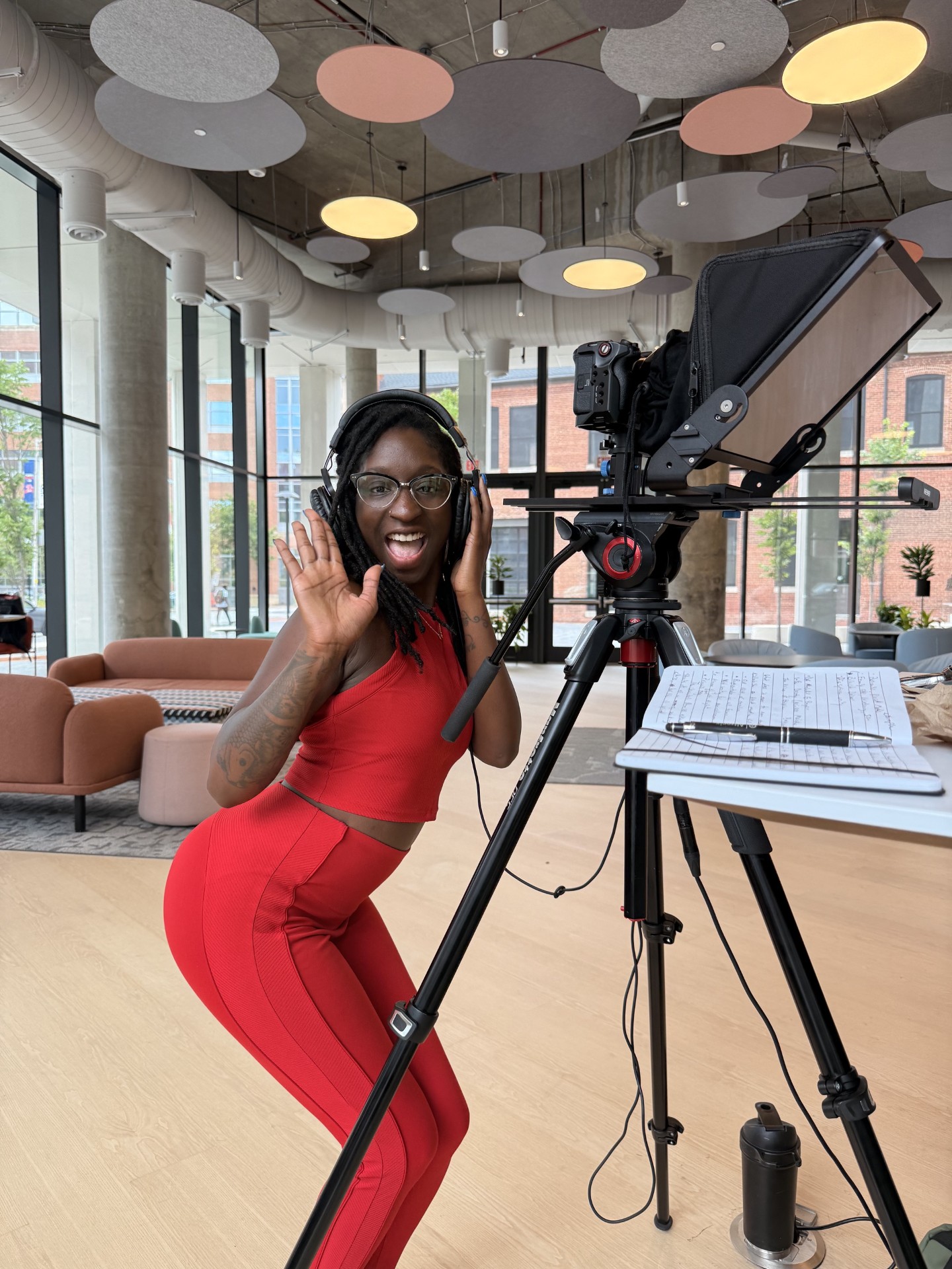A person in red waves beside a camera and teleprompter in a bright studio.