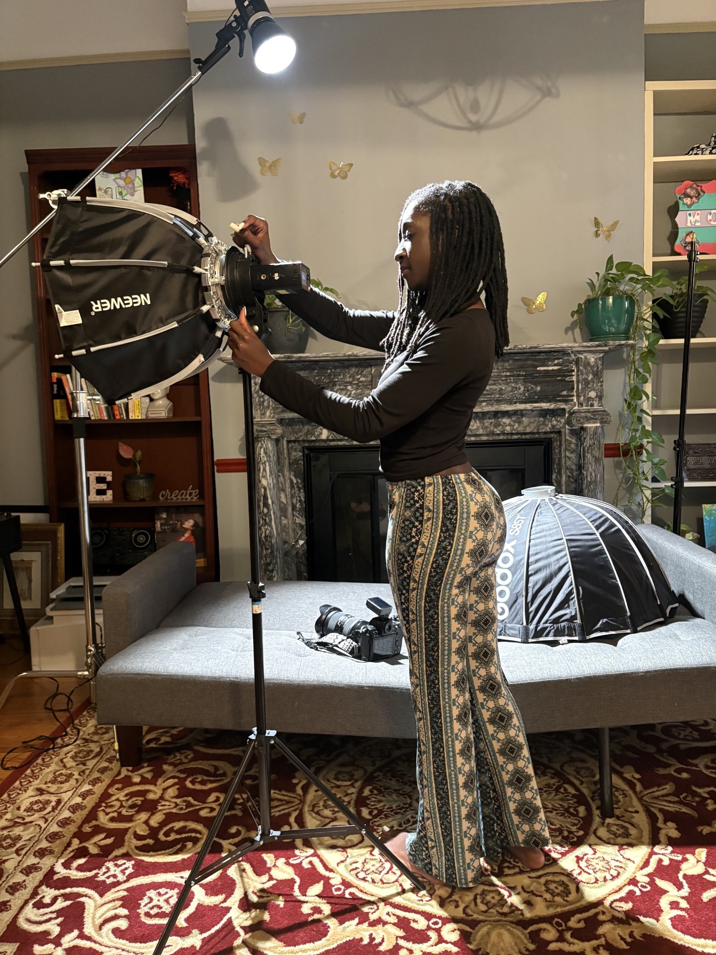 A person adjusts a camera beside a studio light and a bed in a furnished room.