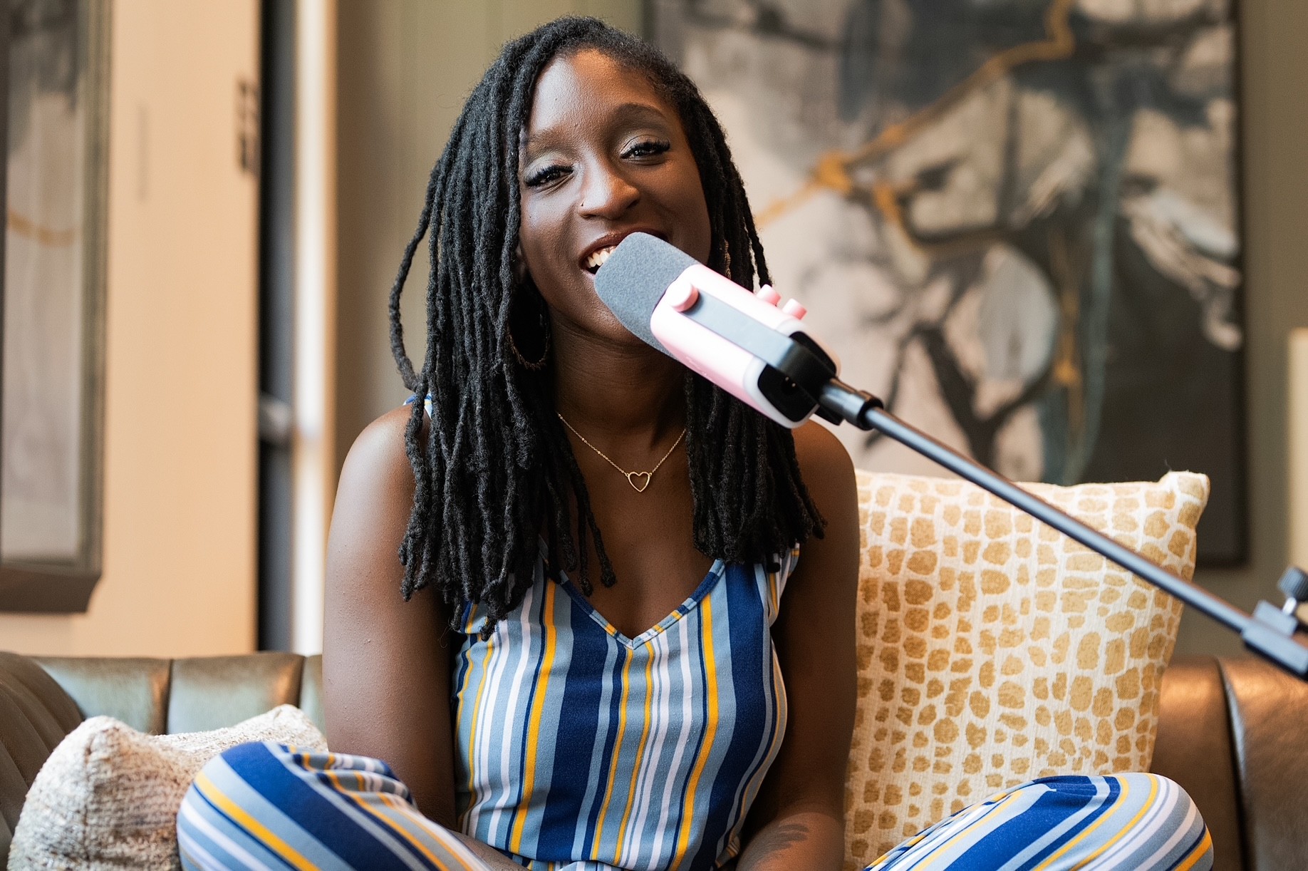 A seated person with long locs holds a microphone, wearing a striped outfit beside patterned cushions.