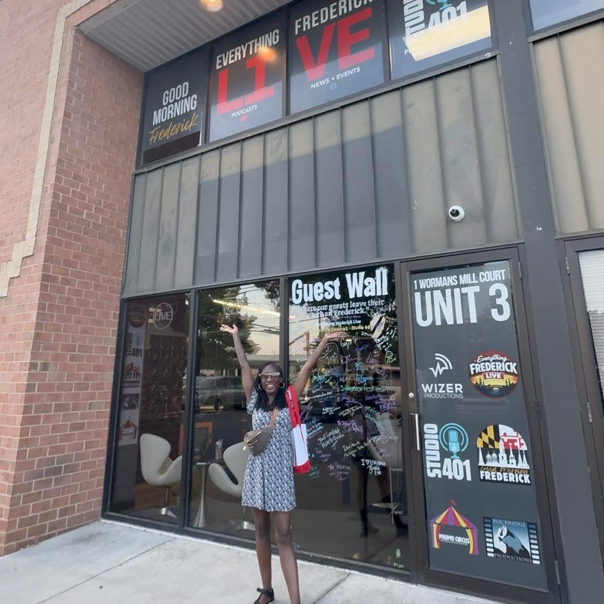 Person standing outside a storefront with window signs and a guest wall.