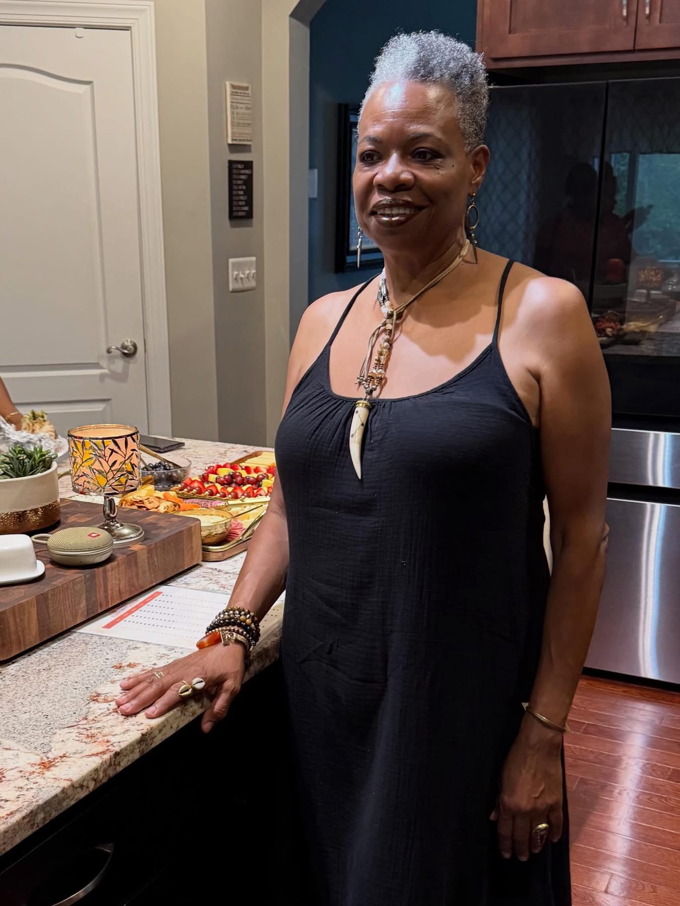 Person standing beside a kitchen counter with food and dishes, wearing a black sleeveless outfit and necklace.